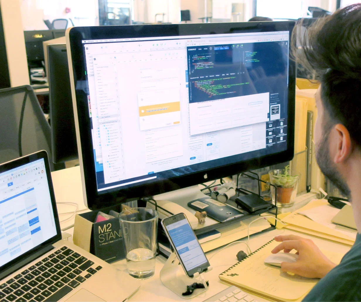 Person working at a desk with multiple screens displaying coding and email applications, surrounded by a laptop, smartphone, glass of water, and office supplies.