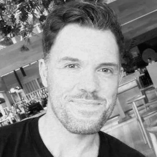 Black and white photo of a smiling man with short hair and a beard, outdoors near trees and a building.