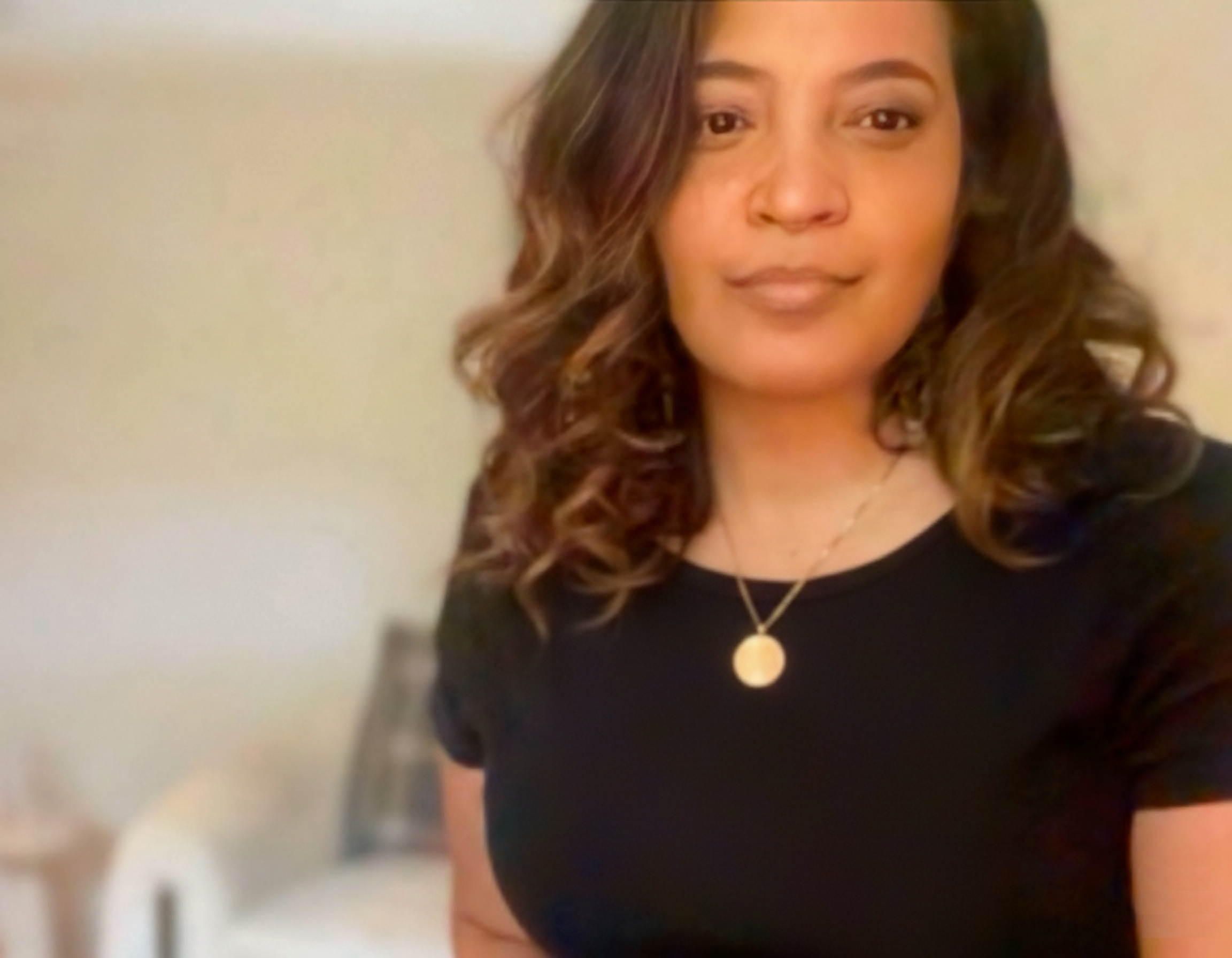 A woman with wavy brown hair wearing a black shirt and a gold necklace taking a selfie indoors.