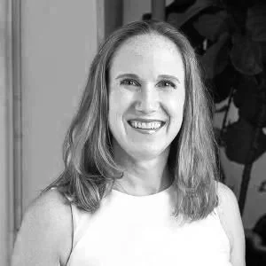 A smiling woman with shoulder-length hair, wearing a sleeveless top, standing indoors with a plant in the background.