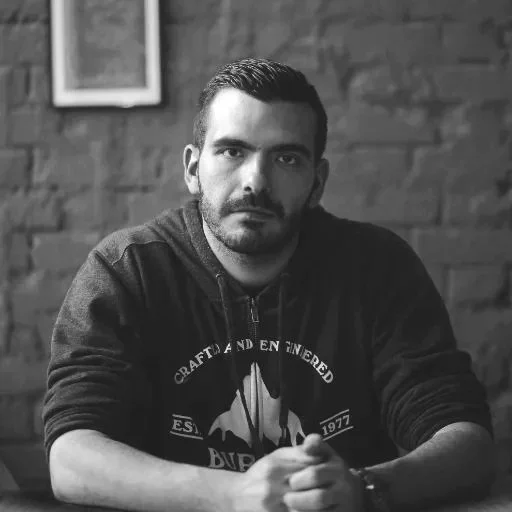 Black and white photo of a man with short dark hair and a beard, wearing a hoodie, sitting at a table in front of a brick wall.