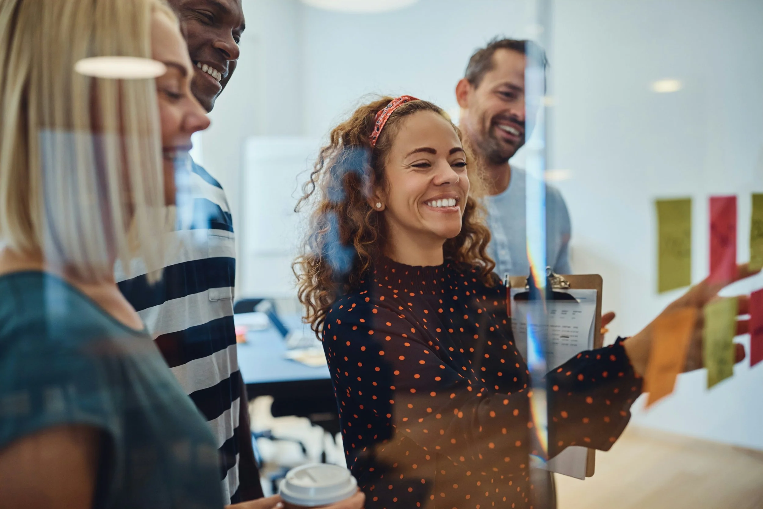 Group of five diverse coworkers smiling while looking at a wall with sticky notes or a whiteboard in an office.
