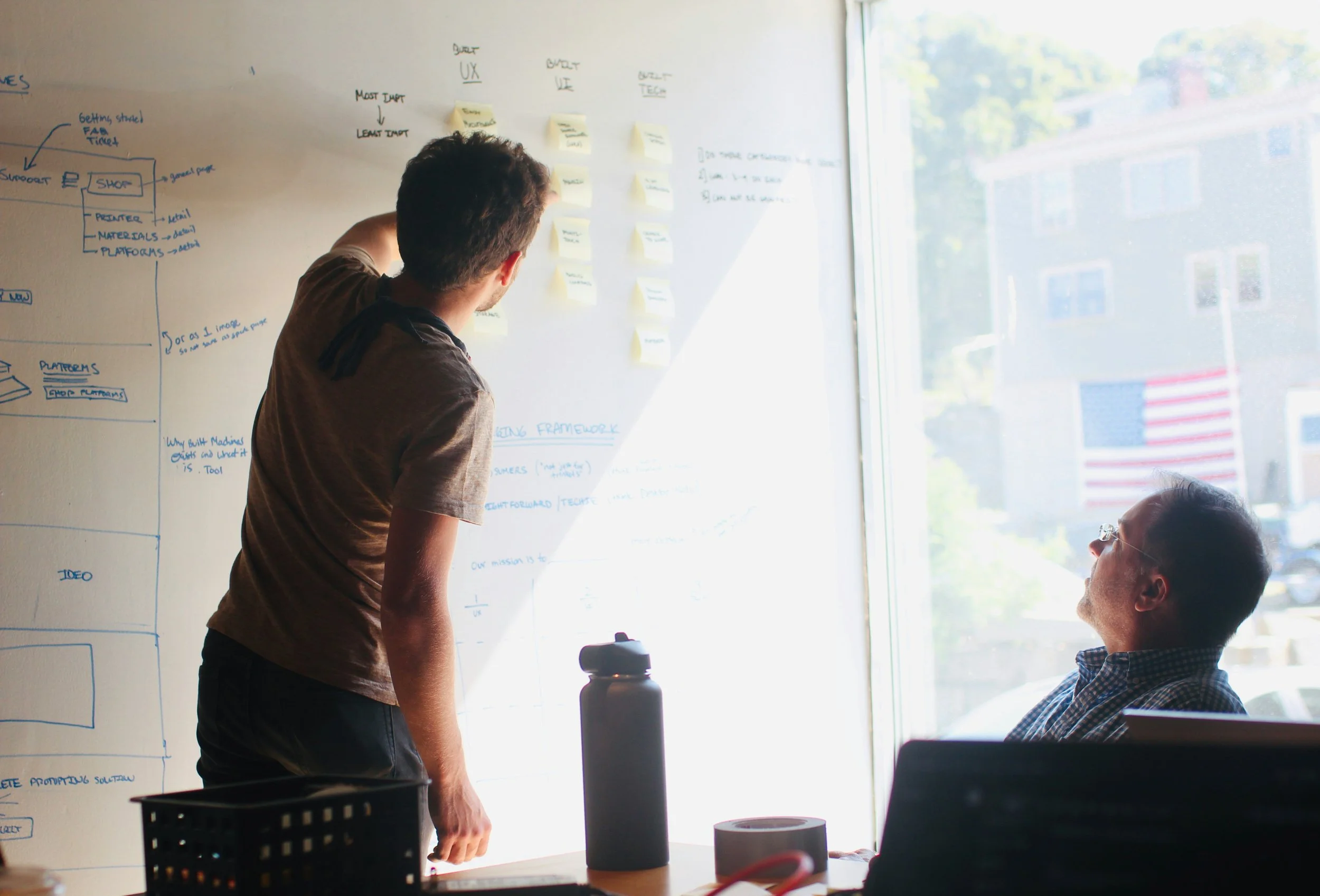 Two men having a conversation in front of a whiteboard with yellow sticky notes and handwritten notes, in a room with large windows revealing a building and trees outside.