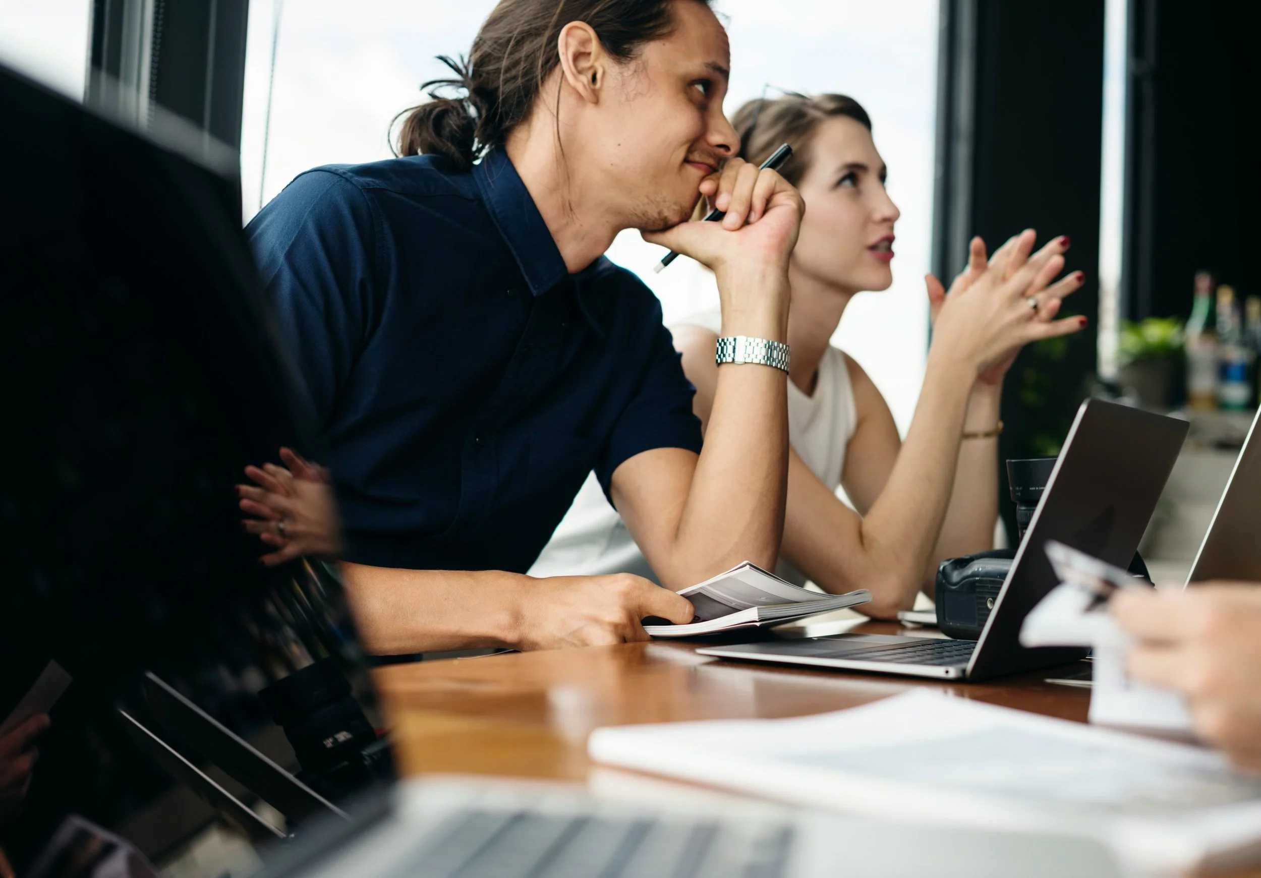 Two people sitting at a table during a meeting, one man in a dark blue shirt holding a pen and looking thoughtful, and a woman with her hands clasped together in conversation.