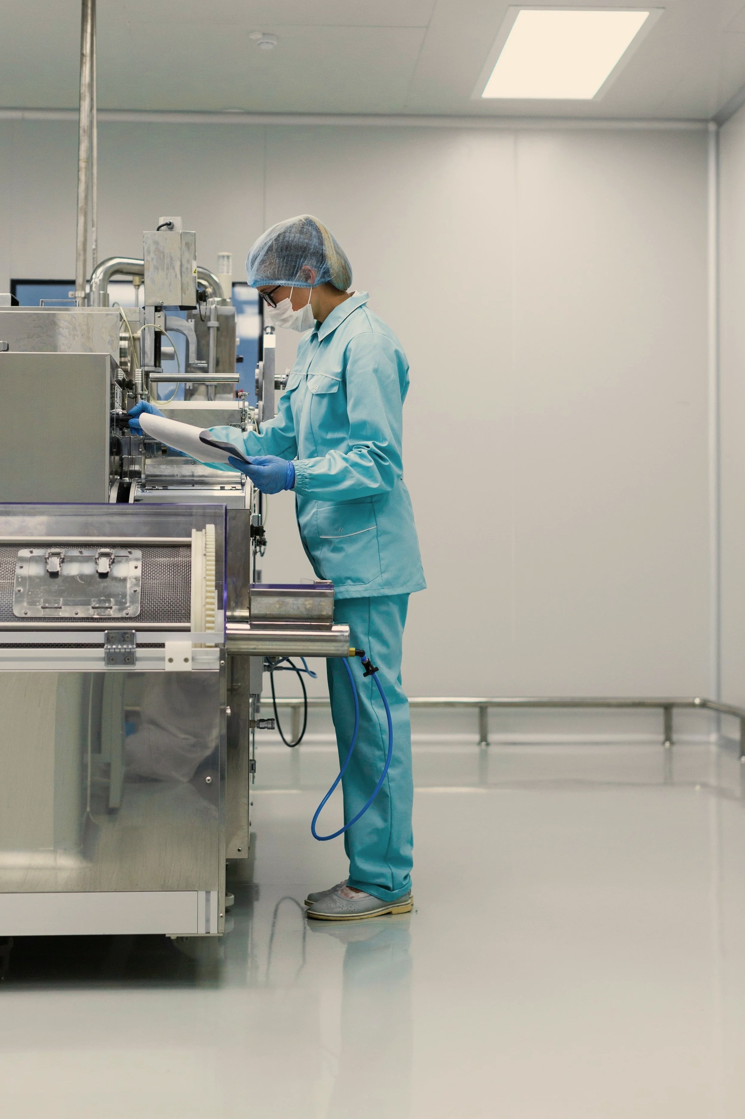 A scientist or technician wearing protective gear, including a hairnet, face mask, glasses, and gloves, working with a modern stainless steel laboratory instrument in a sterile, well-lit lab.