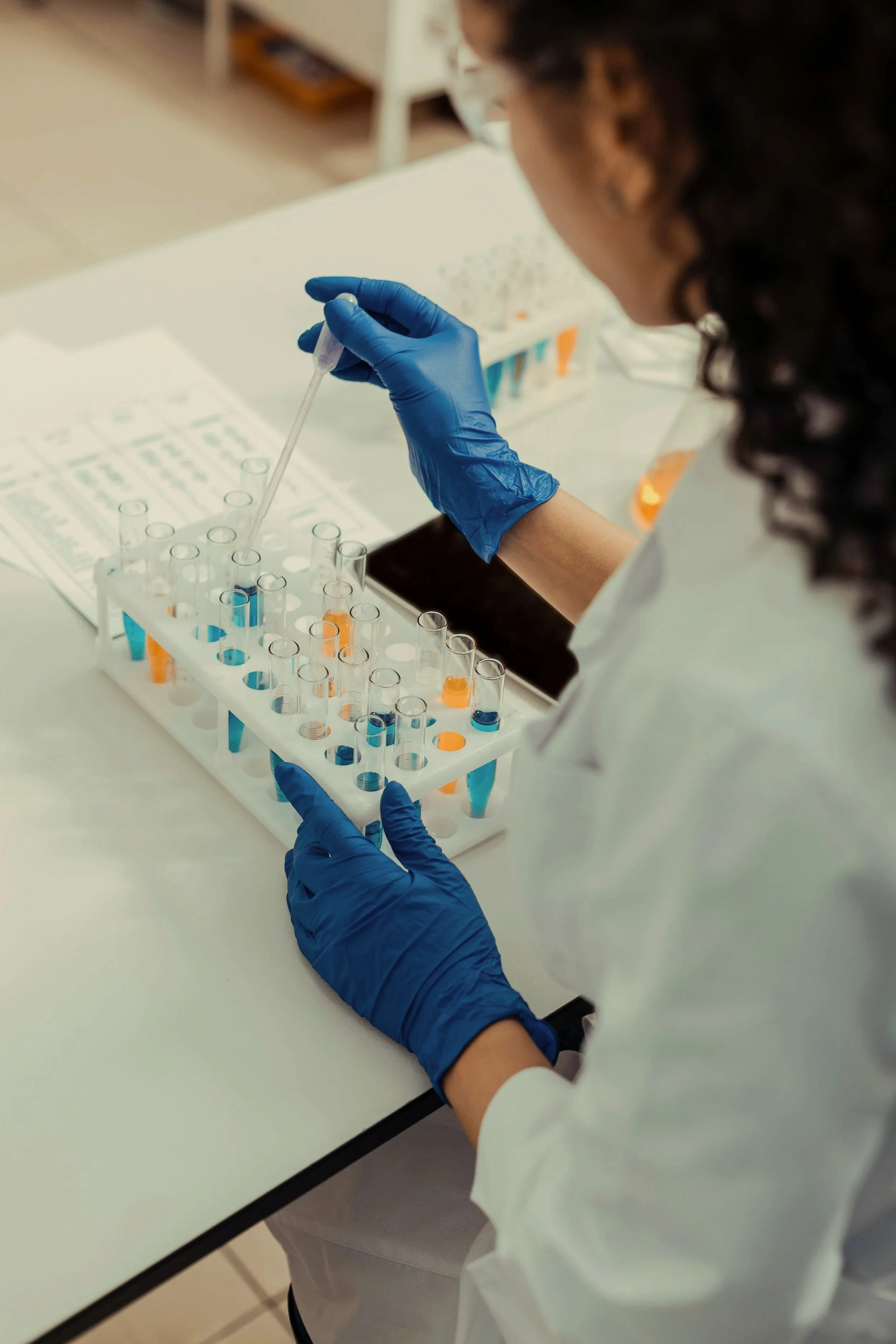 A scientist wearing blue gloves and a white lab coat is working in a laboratory, handling test tubes with blue and orange liquids.