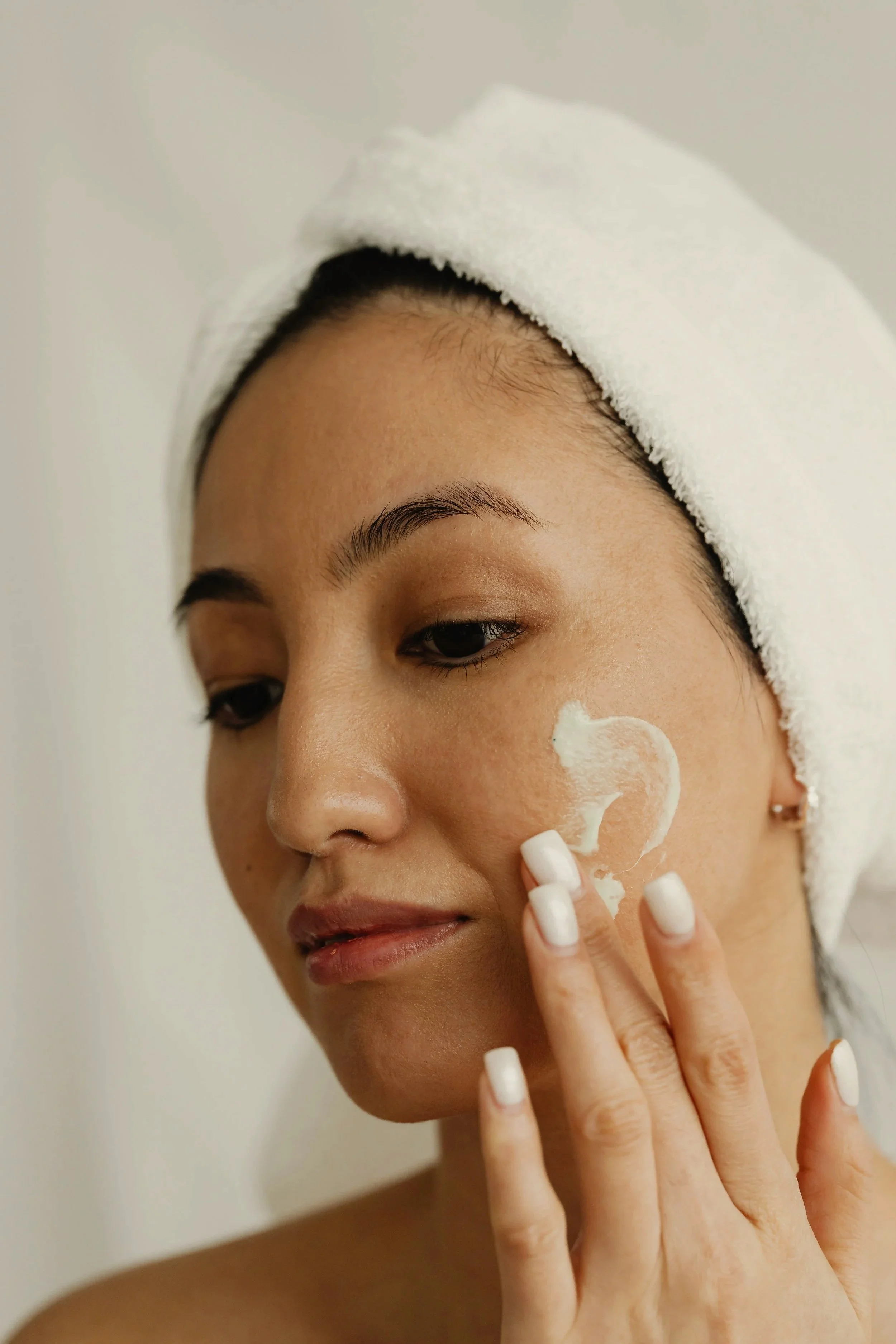 A woman with a white towel wrapped around her head applying skincare cream to her cheek.