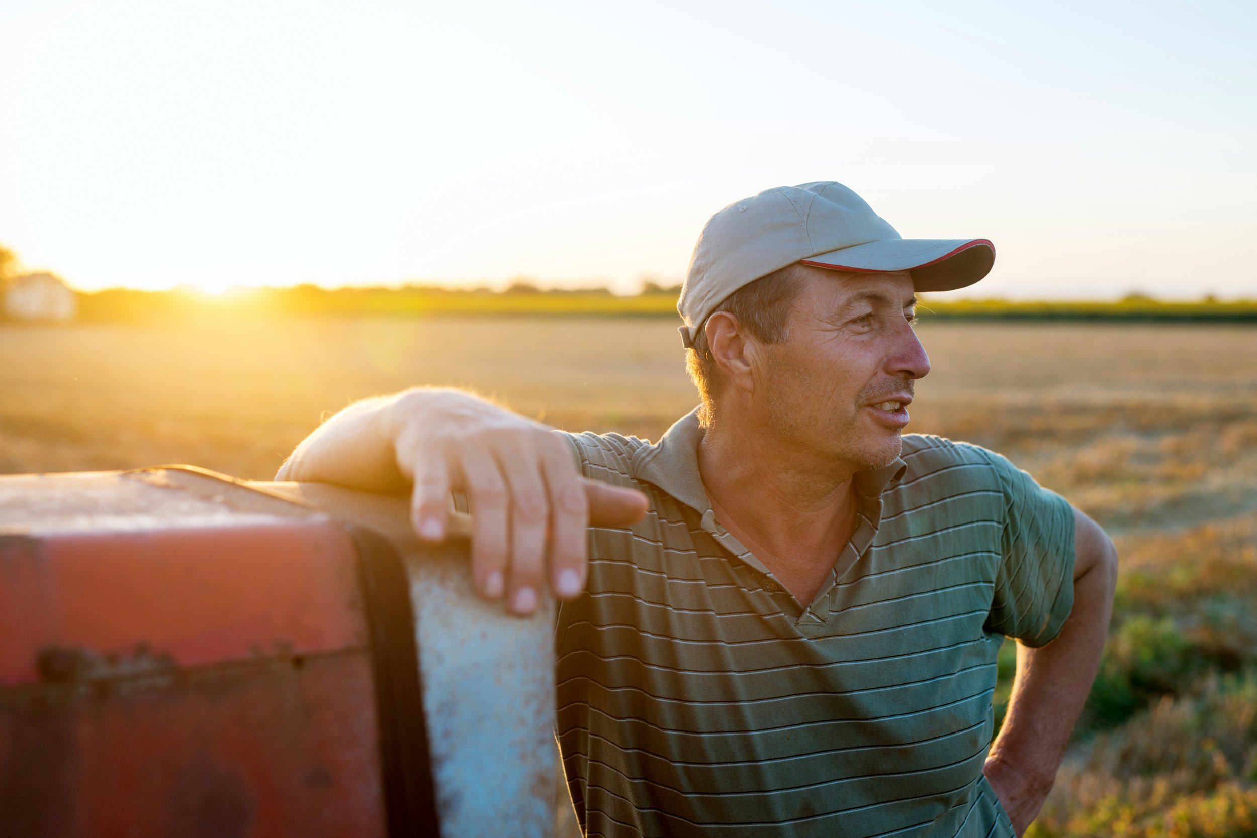 A man wearing a hat and a striped shirt stands outdoors in a field during sunset, resting his arm on a red object.