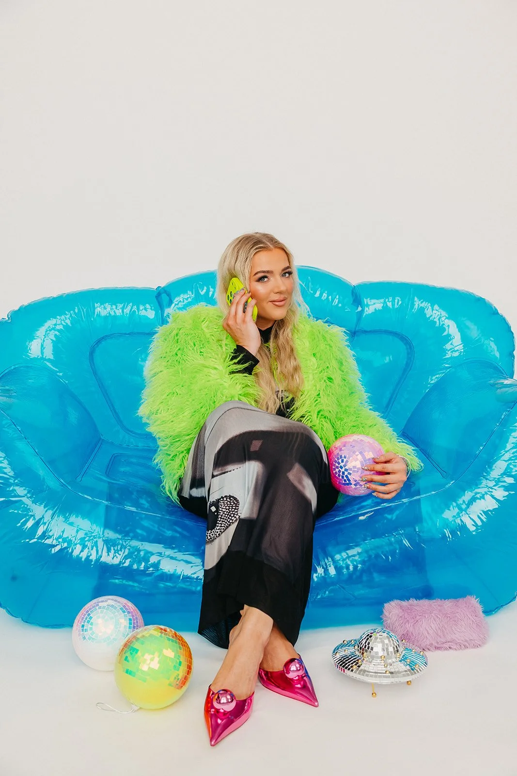 A young woman with long blond hair sitting on a blue inflatable chair, talking on a neon green phone. She is wearing a neon green fuzzy jacket, black and gray pants, and shiny pink high heels. She is holding a pink disco ball, with colorful disco balls and a small pink furry pillow on the floor around her.