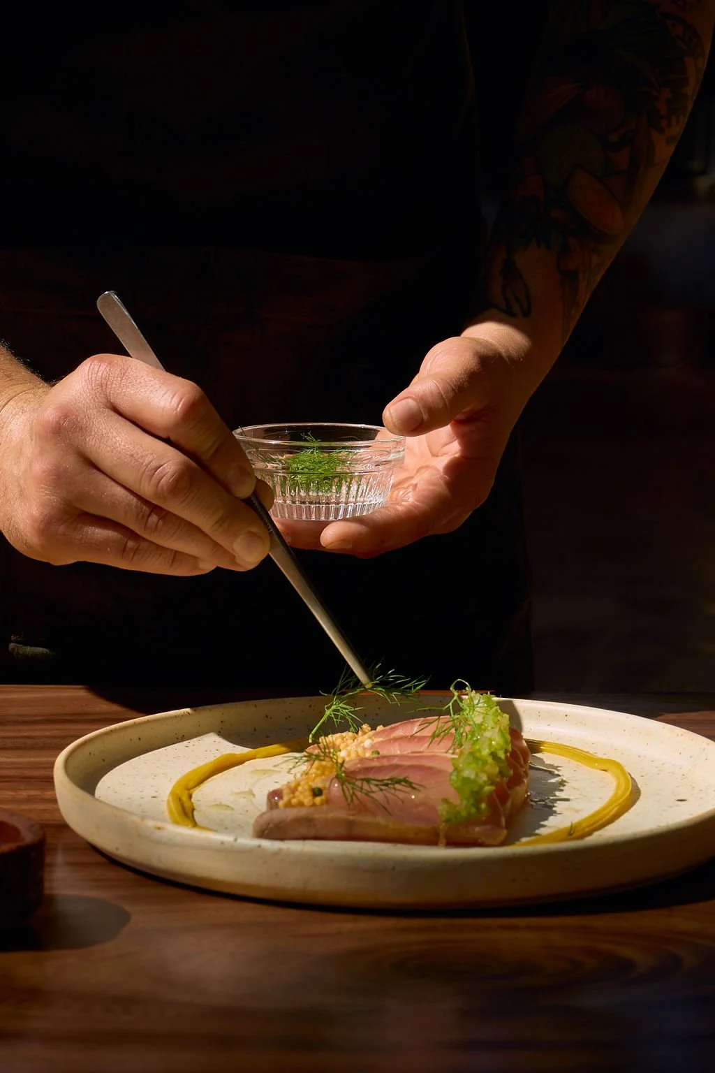 Chef decorando un plato de comida en un plato ovalado con diferentes ingredientes frescos y vegetales, usando una jeringa para añadir detalles.