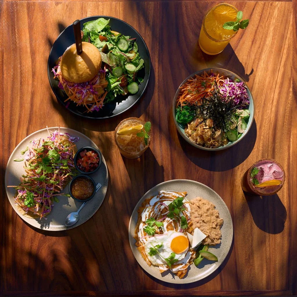 Un restaurante con una variedad de platos que incluyen ensaladas, un sándwich con papas y huevos estrellados, y varios cócteles y bebidas en una mesa de madera.