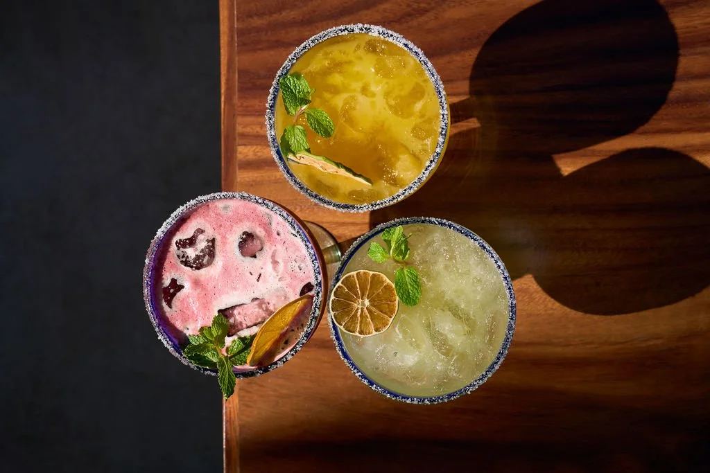 Tres cócteles coloridos con decoraciones de menta, rodajas de limón y otros cítricos en un fondo de madera y sombra, con los bordes escarchados de sal.