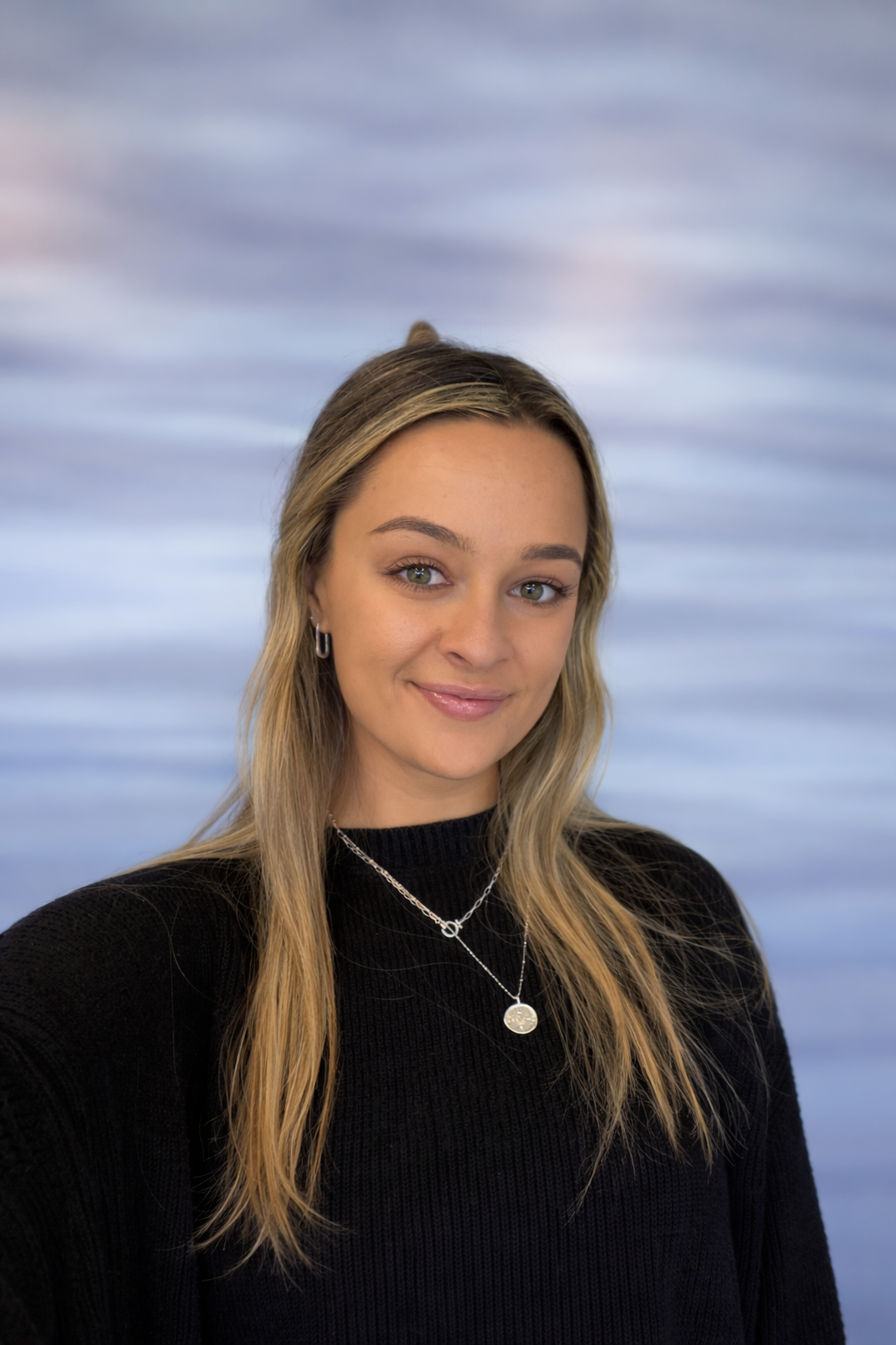 A young woman with long blonde hair wearing a black sweater and jewelry, standing in front of a blue abstract background.