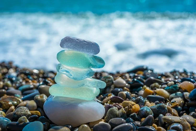 Colorful glass stones stacked on a pebble beach with the ocean in the background.