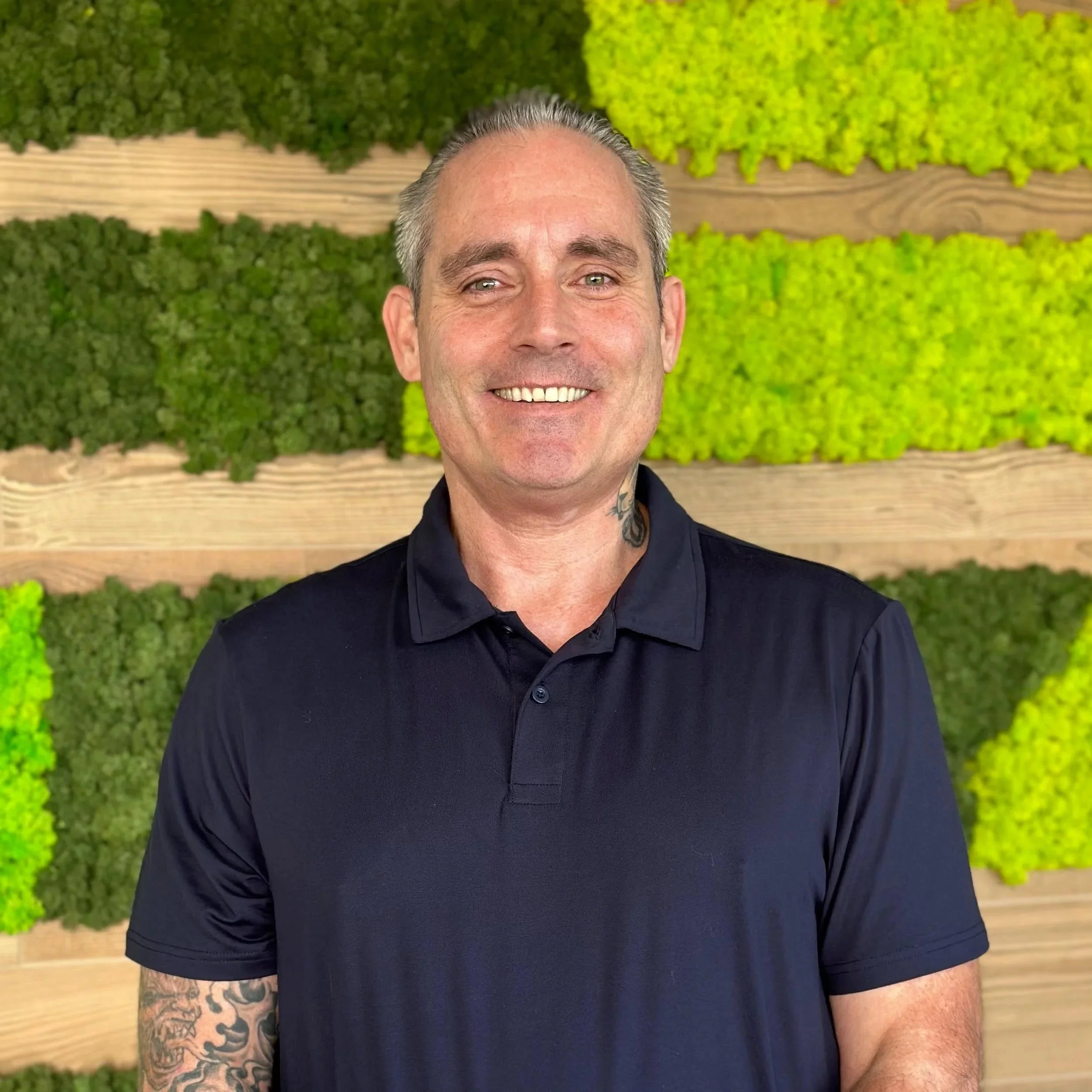 Smiling man in black polo shirt standing in front of a green moss wall with horizontal wooden panels.