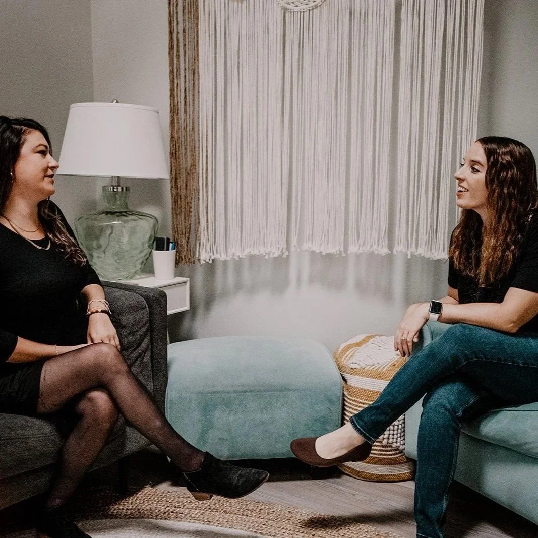Two women sitting in a living room having a conversation, facing each other, with a lamp and a curtain behind them.