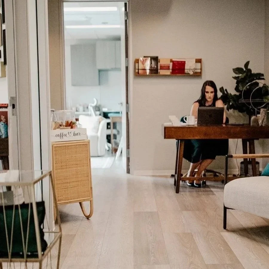 A woman working on a laptop at a wooden desk in a cozy, modern coffee shop or cafe. There is a plant beside her and a small bookshelf on the wall.