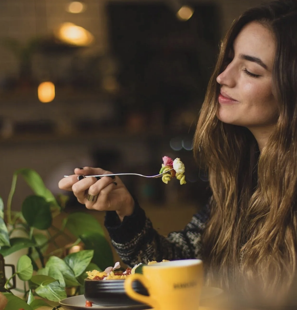 A young woman with long brown hair and a black patterned sweater is eating dessert with a fork. She appears to be enjoying her meal, with closed eyes and a slight smile. There are green plants and a yellow mug on the table, with a blurred background of warm lights, suggesting a cozy indoor setting at night.