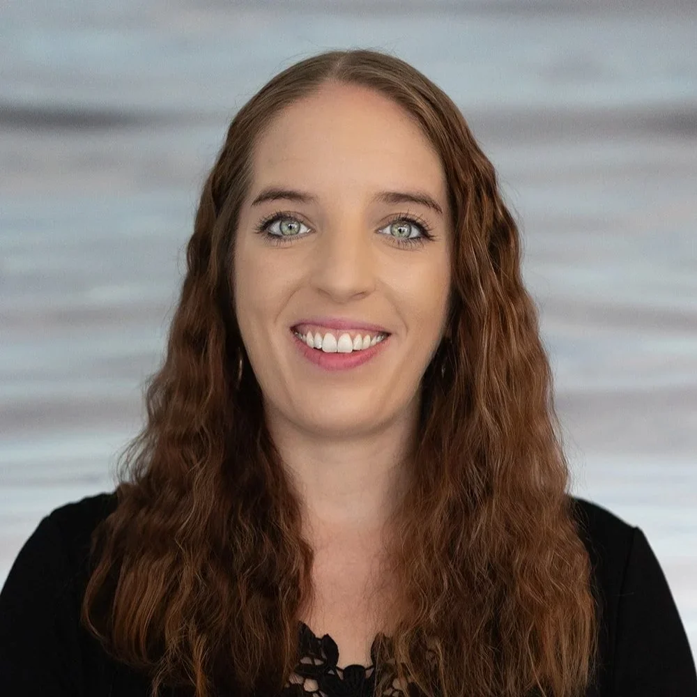 A woman with long, wavy red hair, blue eyes, and a bright smile, wearing a black top, standing in front of a softly blurred blue and gray background.