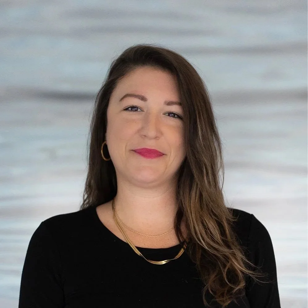 A woman with long brown hair, wearing a black top and gold jewelry, smiling in front of a neutral background.