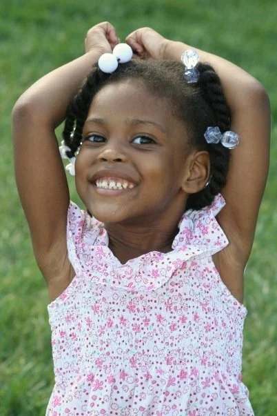 Young girl smiling outdoors, wearing a pink and white floral dress, with her hands on her head, showing her braided hair with beads.