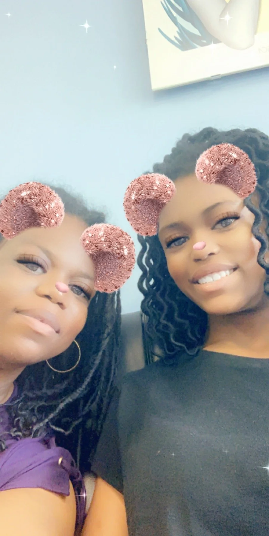 Two women taking a selfie together with a filter that adds pink, fuzzy bear ears and a small pink nose to their faces, both smiling and sitting close to each other in an indoor setting.