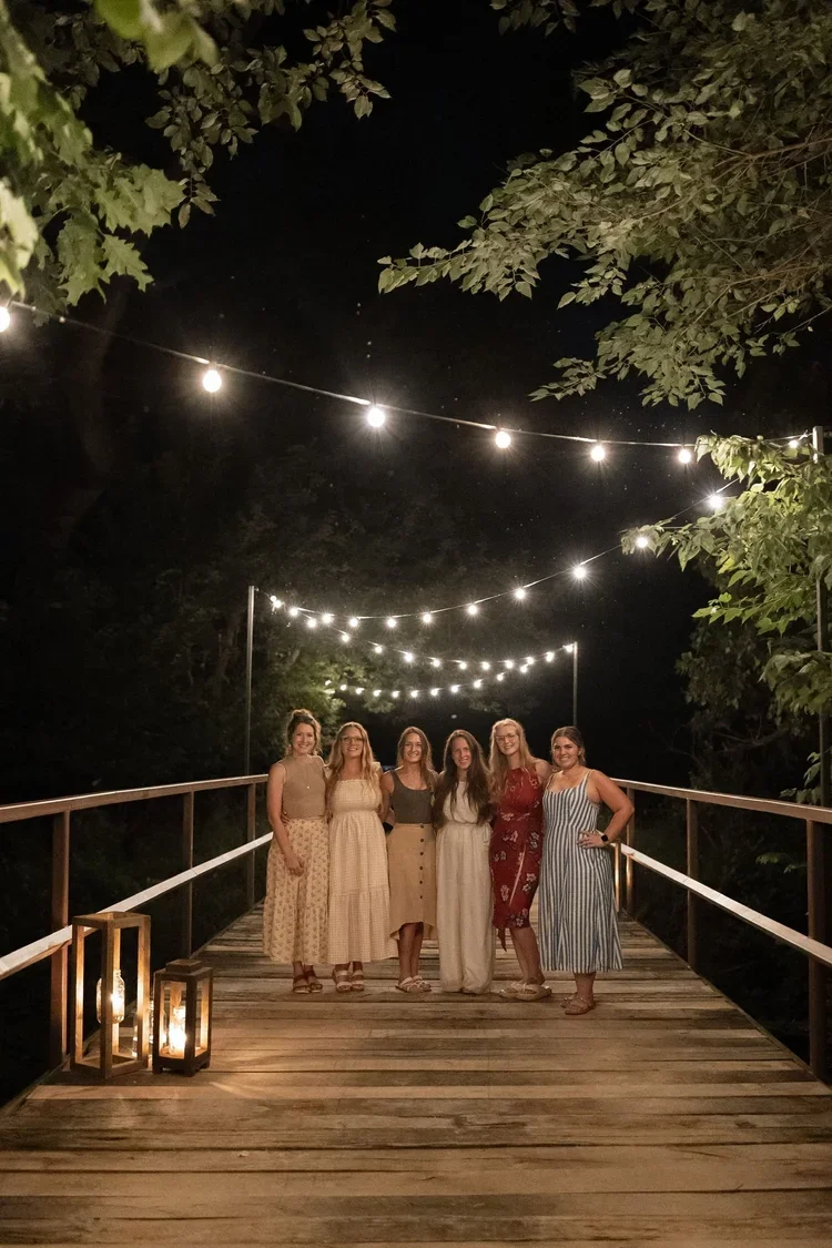 Five women standing on a wooden bridge at night, surrounded by string lights and trees.