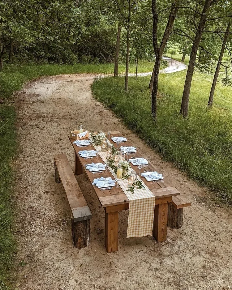 A rustic outdoor dining table set on a dirt path in a wooded area, decorated with a yellow checkered table runner, white napkins, glasses, candles, and greenery.