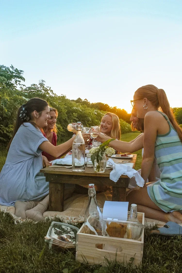 A group of women enjoying a picnic outdoors at sunset, sitting around a low wooden table with drinks and food, surrounded by elderflowers.