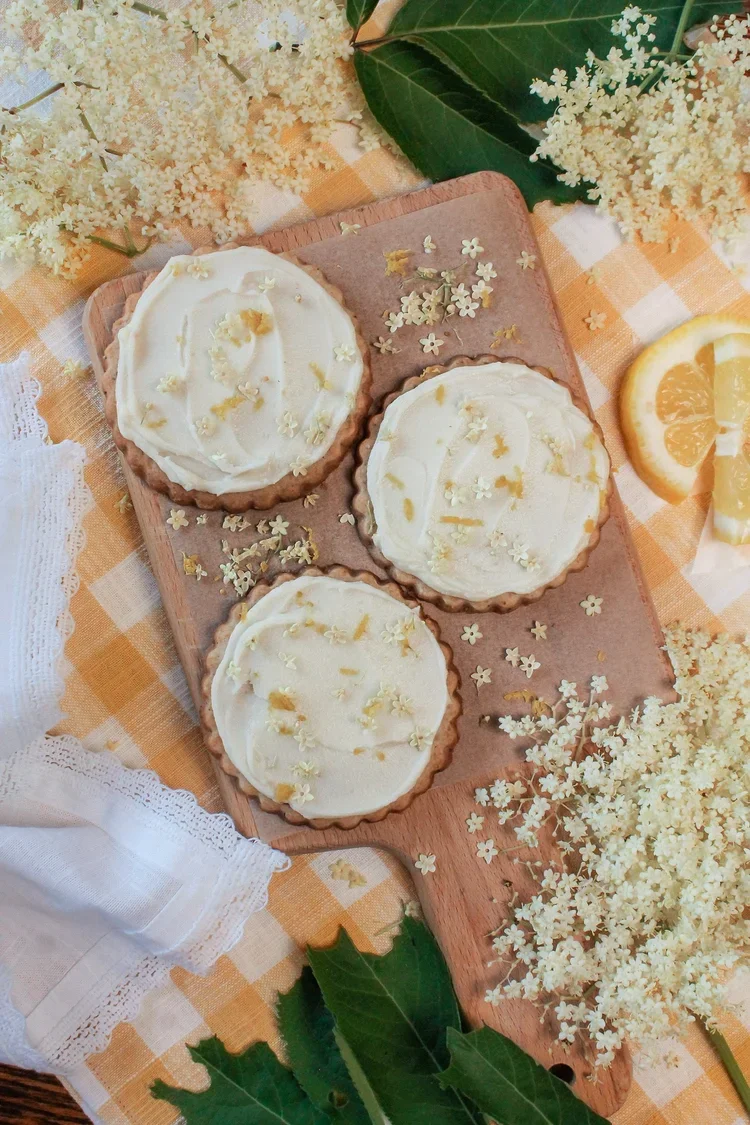 Elderflower shortbread cookies with white icing decorated with small lemon and freeze-dried elderflowers on a wooden board surrounded by white flowers, lemon slices, green leaves, and a checkered tablecloth.