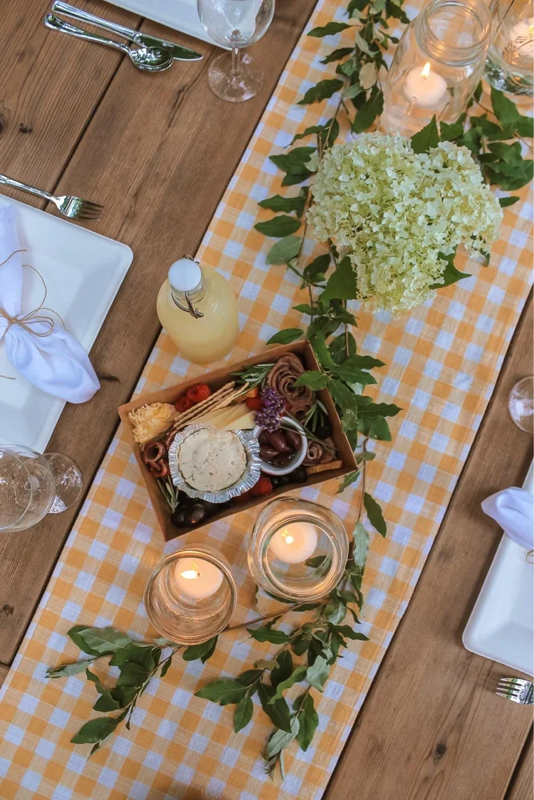 A decorated dining table with a yellow checkered table runner, green foliage, white hydrangea centerpiece, candles in glass jars, a bottle of lemonade, white plates with napkins, and a cheese and charcuterie platter.