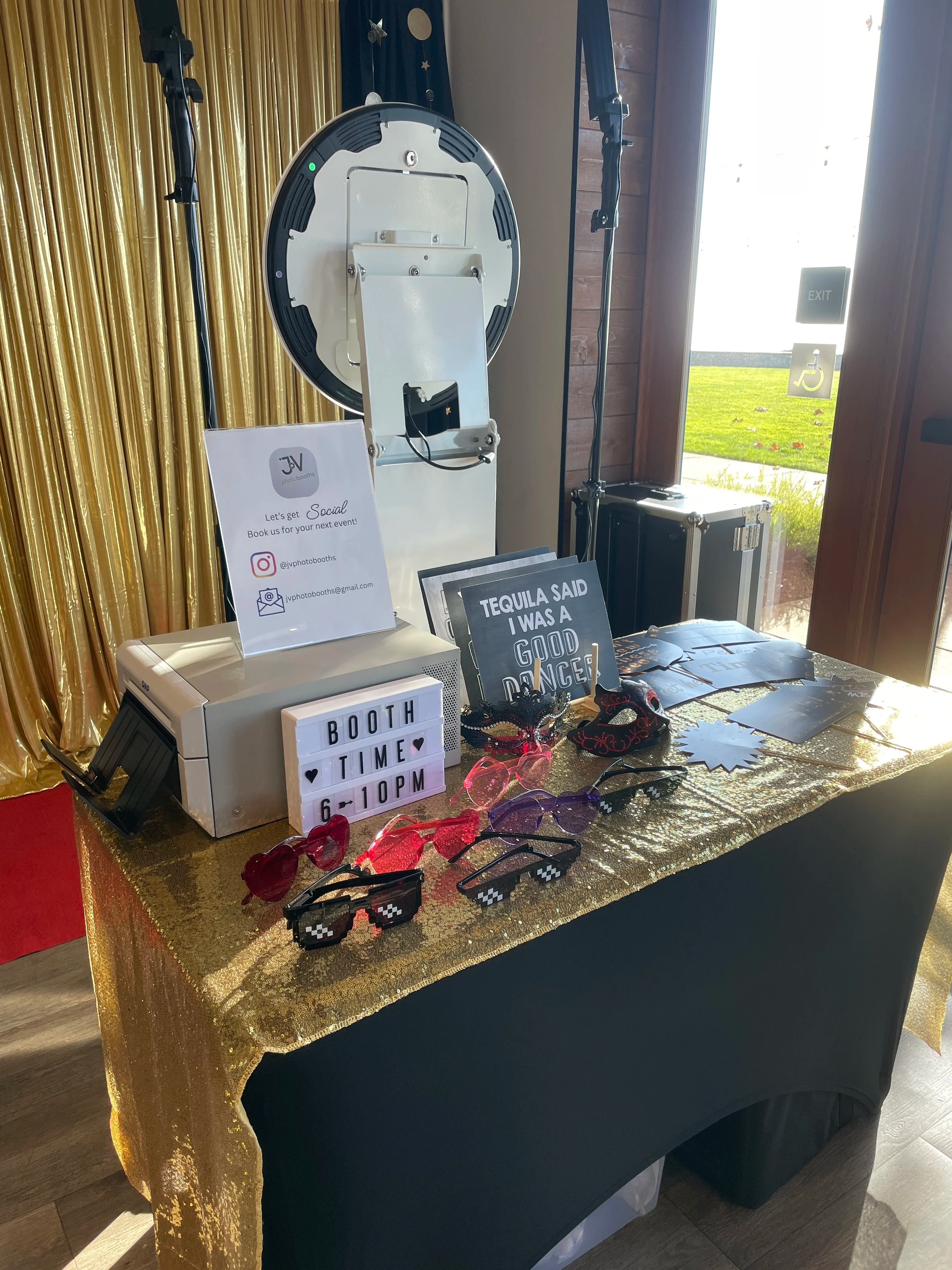 Photo of a photo booth setup with a gold cloth cover, sunglasses, sign that says 'Tequila Said I Was a Good Dancer,' and a sign with event details, outside view of green grass through large window.