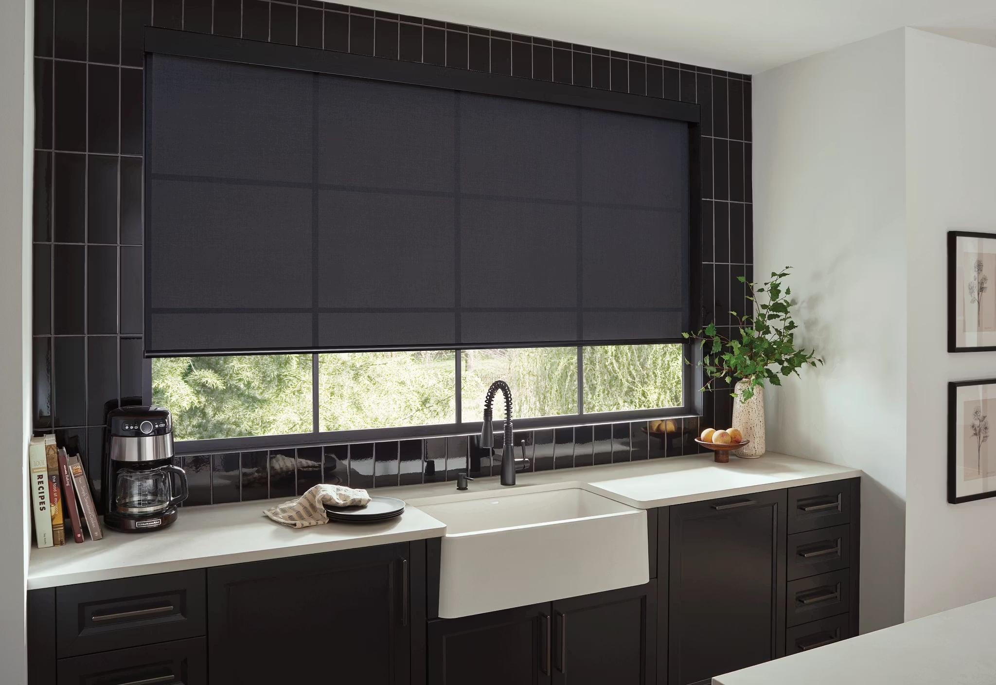 Modern kitchen with black cabinetry, white countertop, black tiled backsplash, and a window with a black roller shade. There is a coffee maker, books, and plates on the counter, a potted plant, and framed wall art.