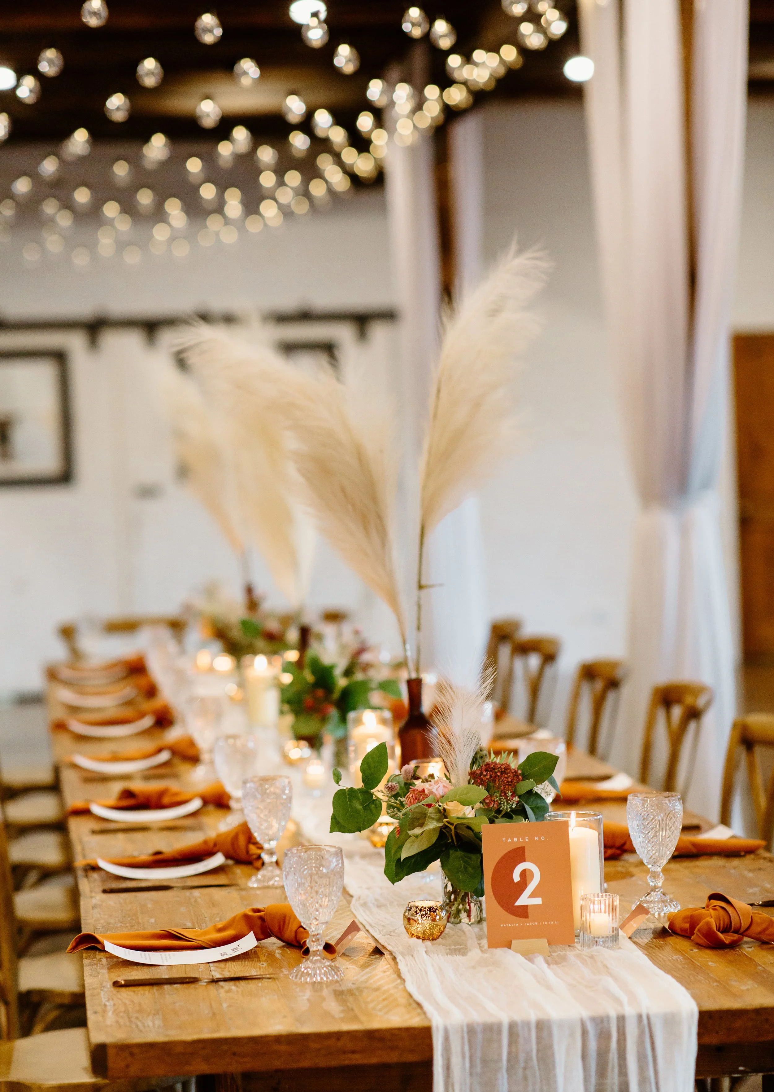 Elegant banquet table decorated with candles, greenery, tall pampas grass, and orange table numbers in a rustic venue with string lights and white curtains.
