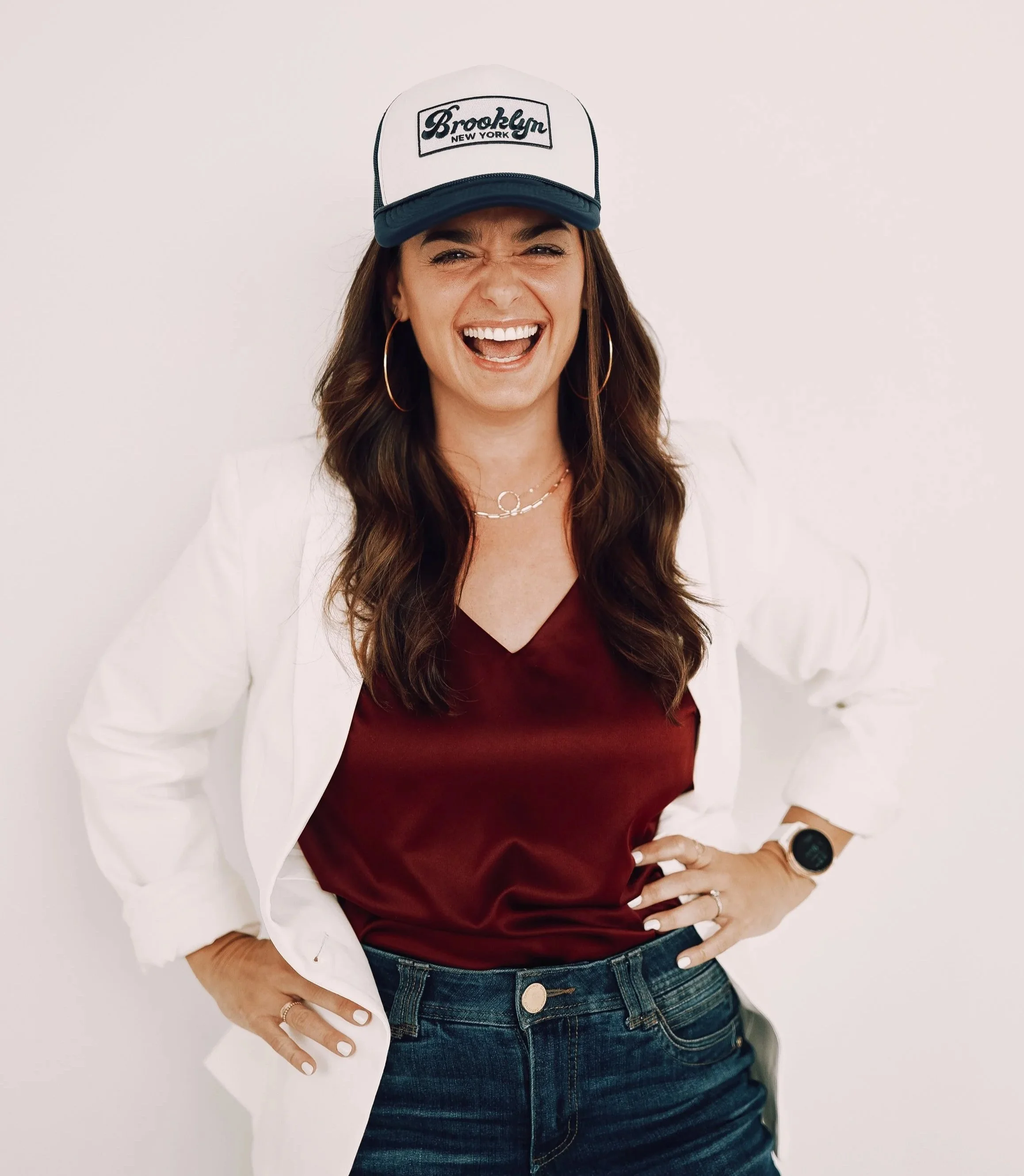 Woman smiling in a white blazer, maroon top, wearing a Brooklyn New York cap, standing against a white background.
