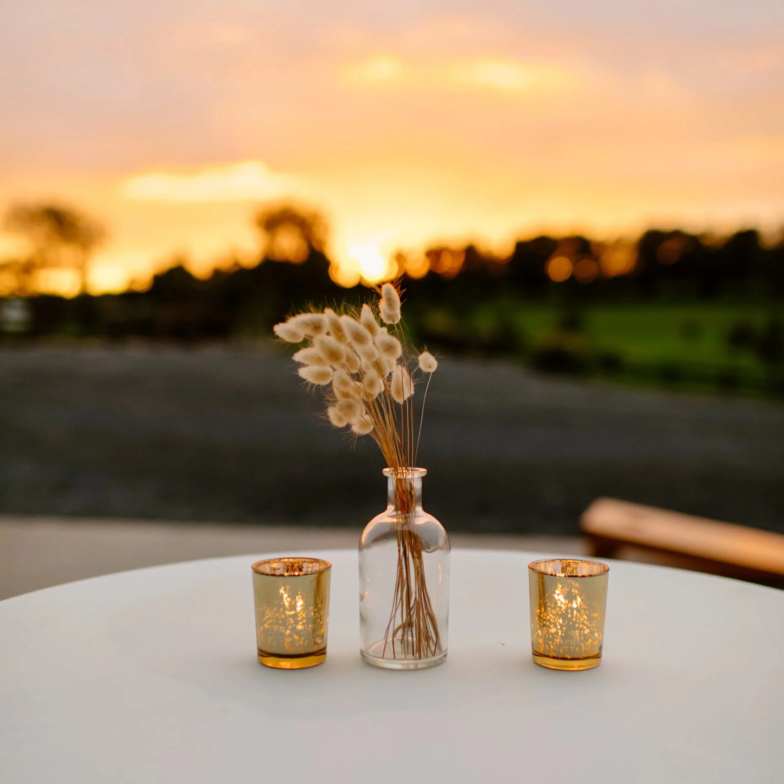 A table with a small clear glass vase containing dried white flowers and two small candle holders with lit candles, set outdoors against a blurred sunset sky.