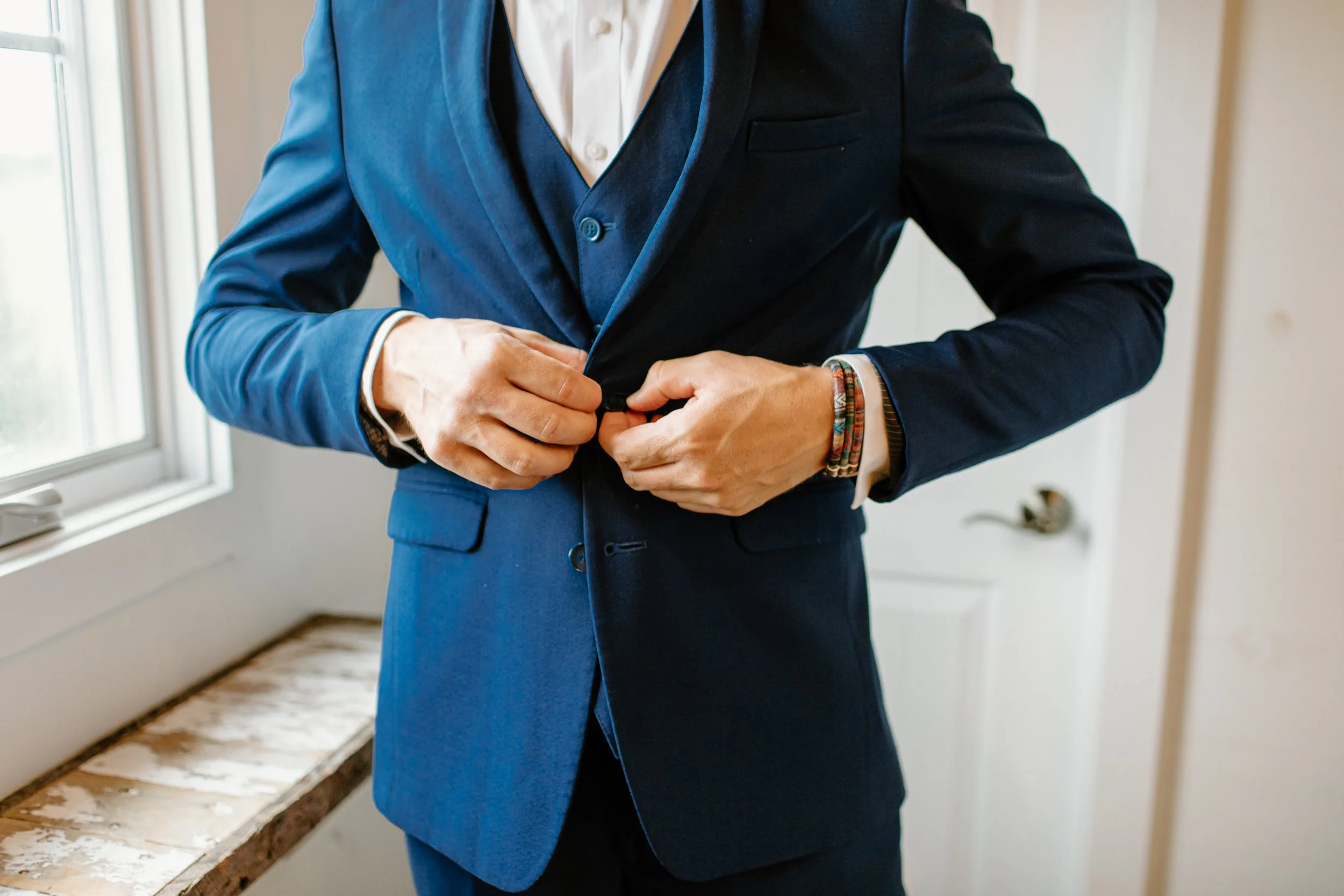 Person in a navy blue suit buttoning the jacket near a window.