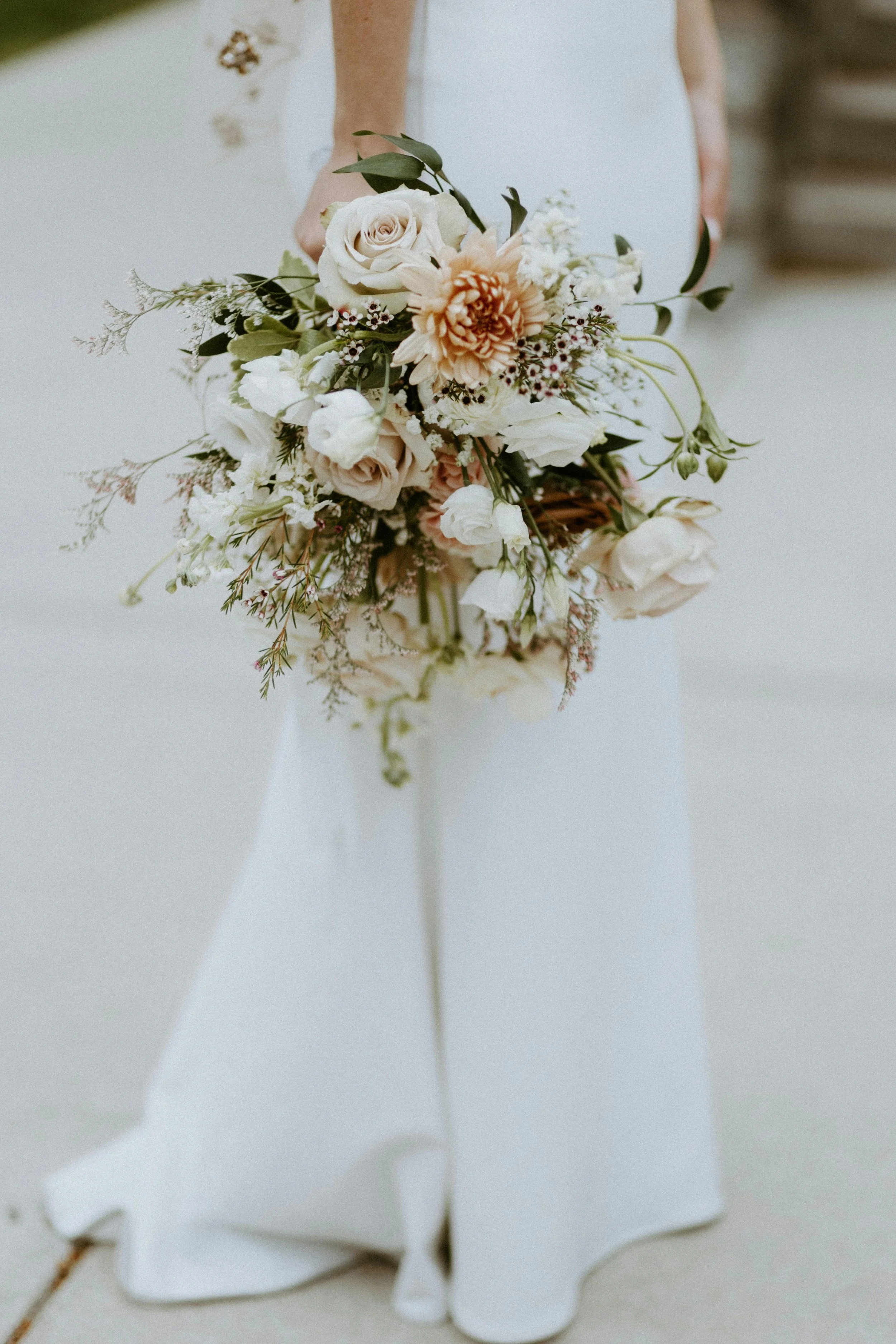A person in a white dress holding a bouquet of flowers, including roses and peonies, with a blurry outdoor background.