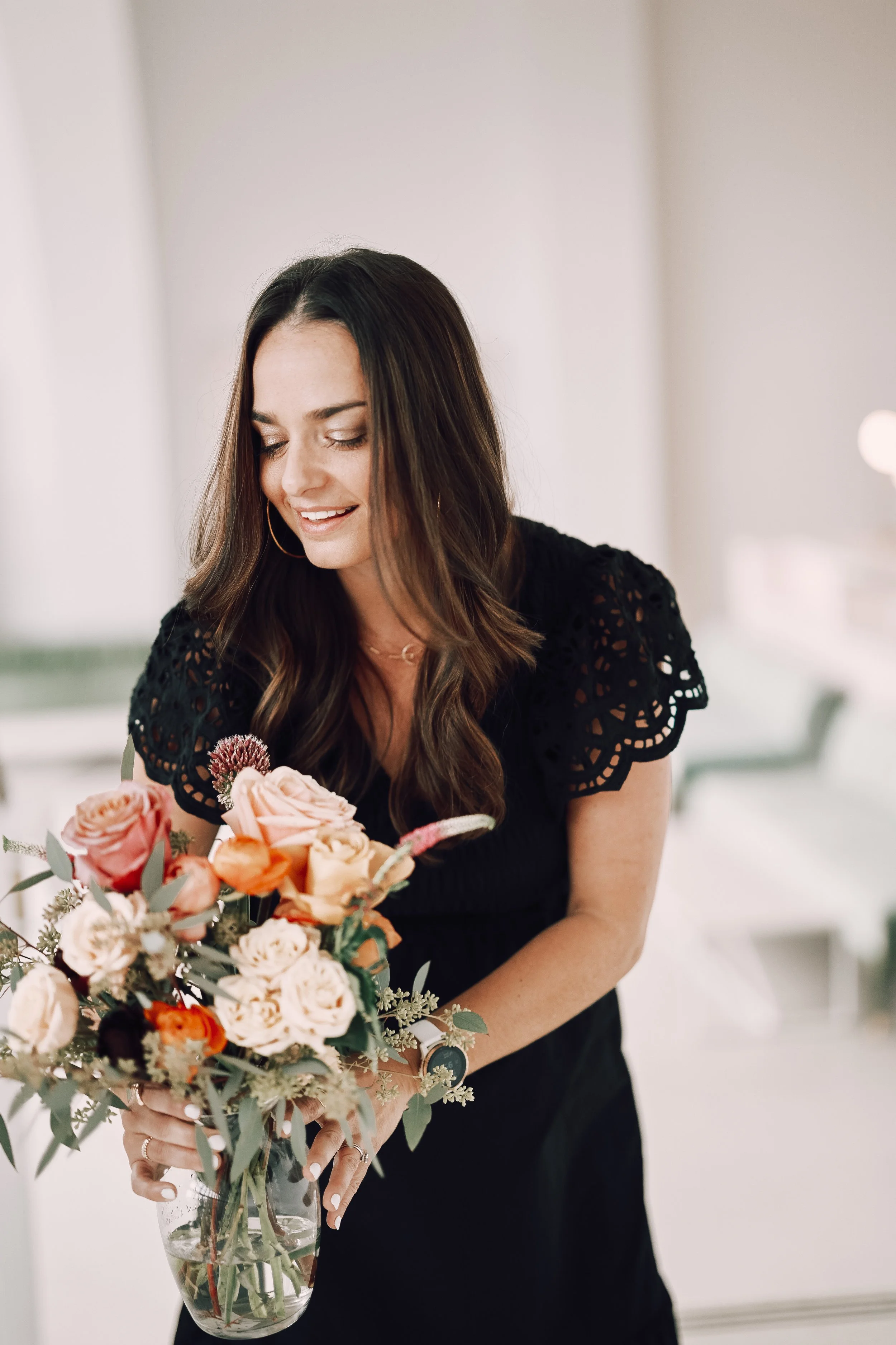 A woman with medium-length brown hair wearing a black dress with lace sleeves stands smiling while holding a bouquet of pink, peach, and cream flowers in a glass vase.