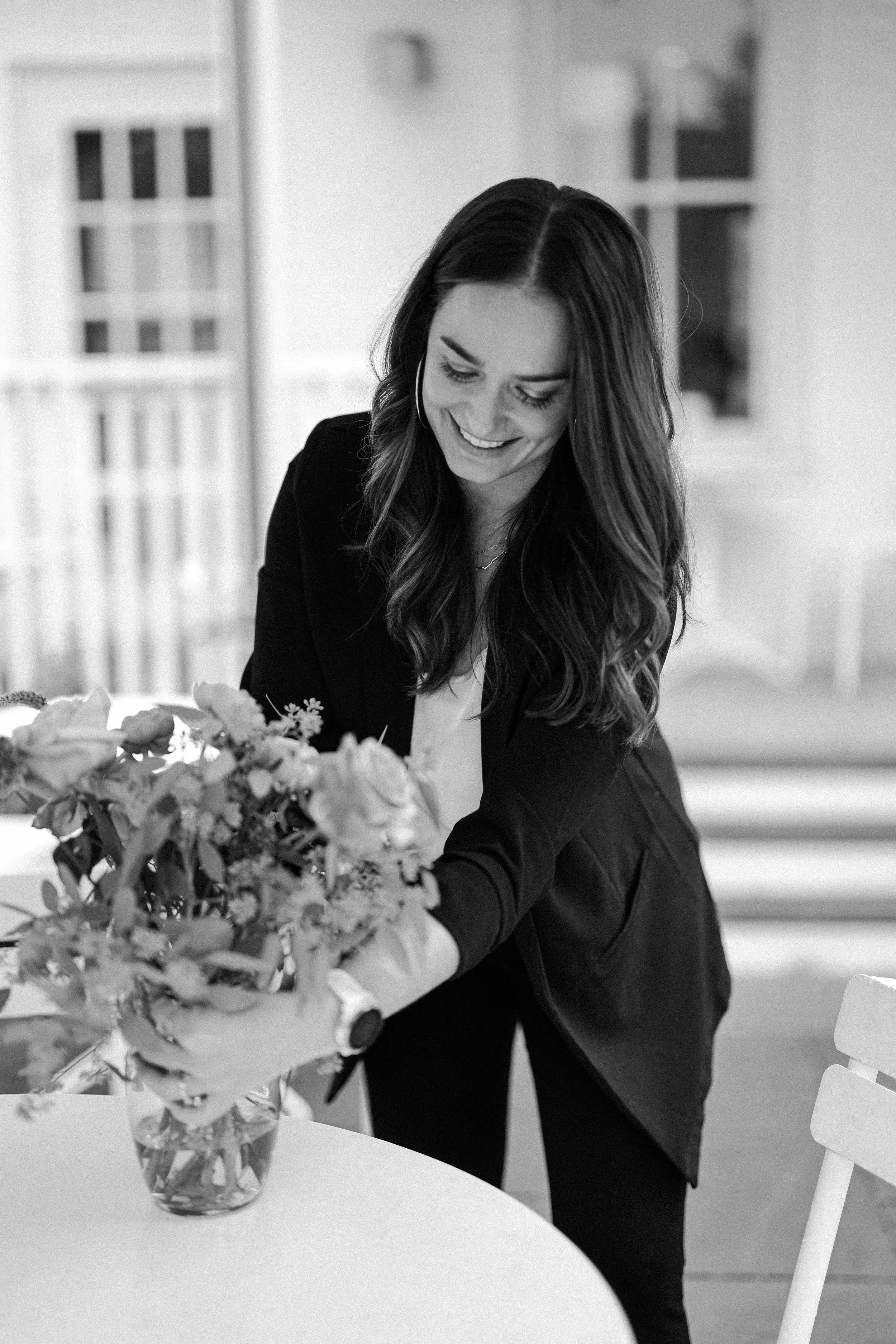 A woman with long wavy hair, smiling, arranging a bouquet of flowers in a glass vase on a table.