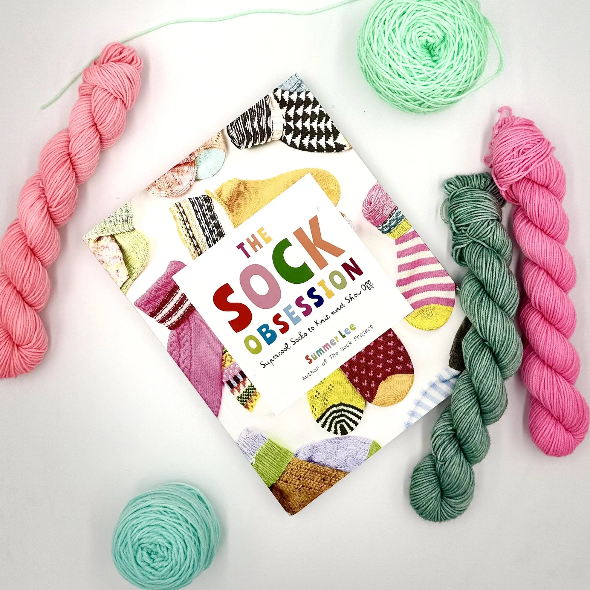 Book titled 'The Sock Obsession' surrounded by colorful skeins of yarn and a fabric with sock patterns.
