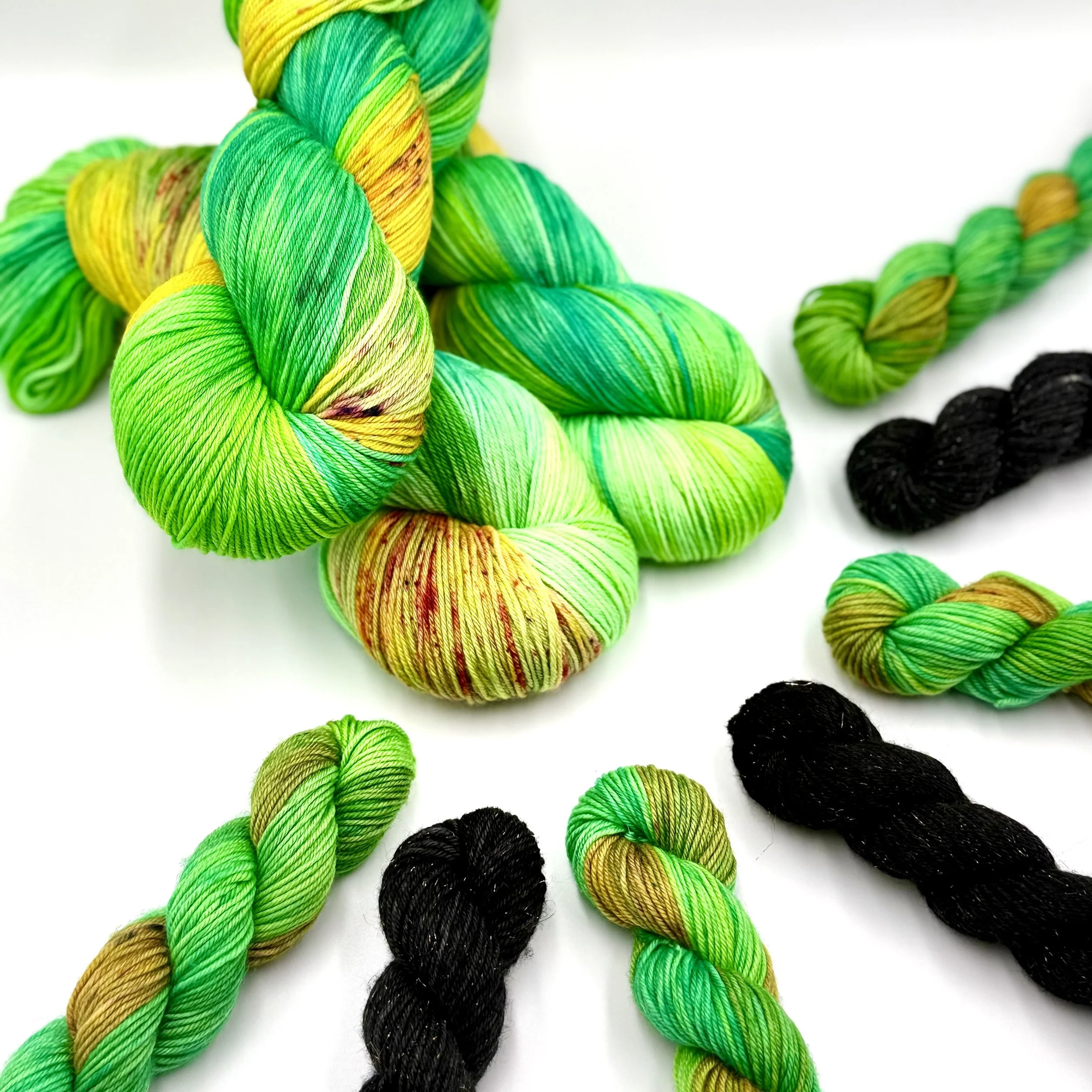 Colorful yarn skeins hand dyed in various shades of green, black, and a mix of yellow and brown, arranged on a white background.