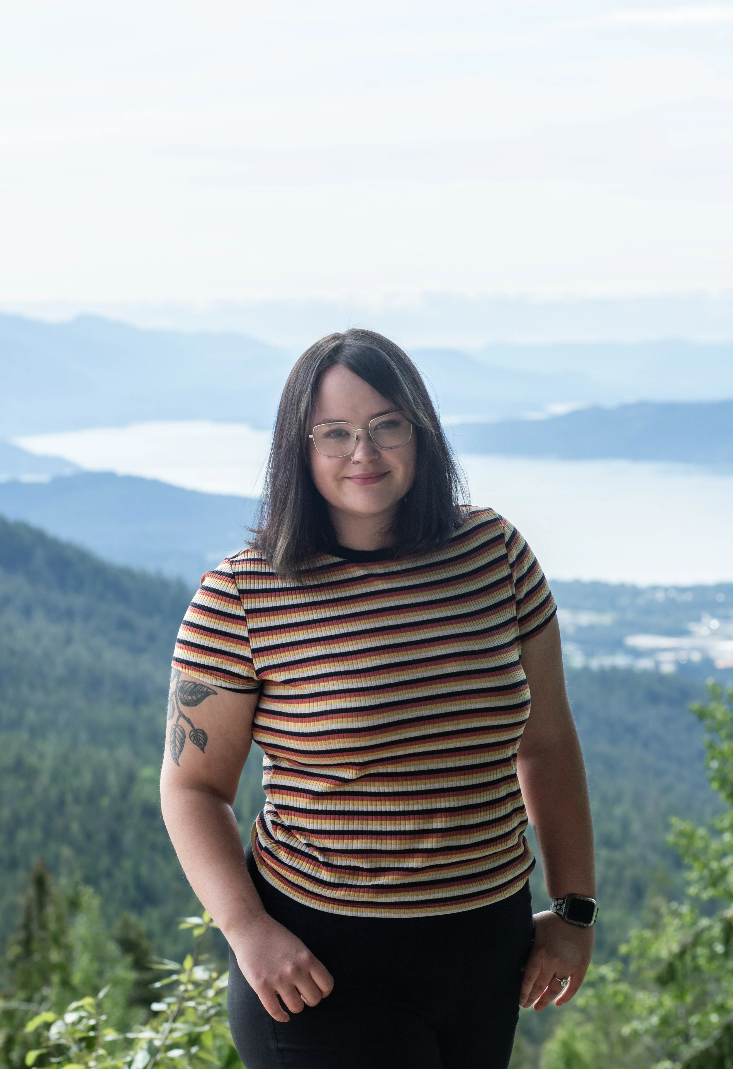 A woman with shoulder-length dark hair, glasses, and a tattoo on her left arm, standing outdoors in front of a scenic mountain and lake view. She is wearing a striped T-shirt and a smartwatch.