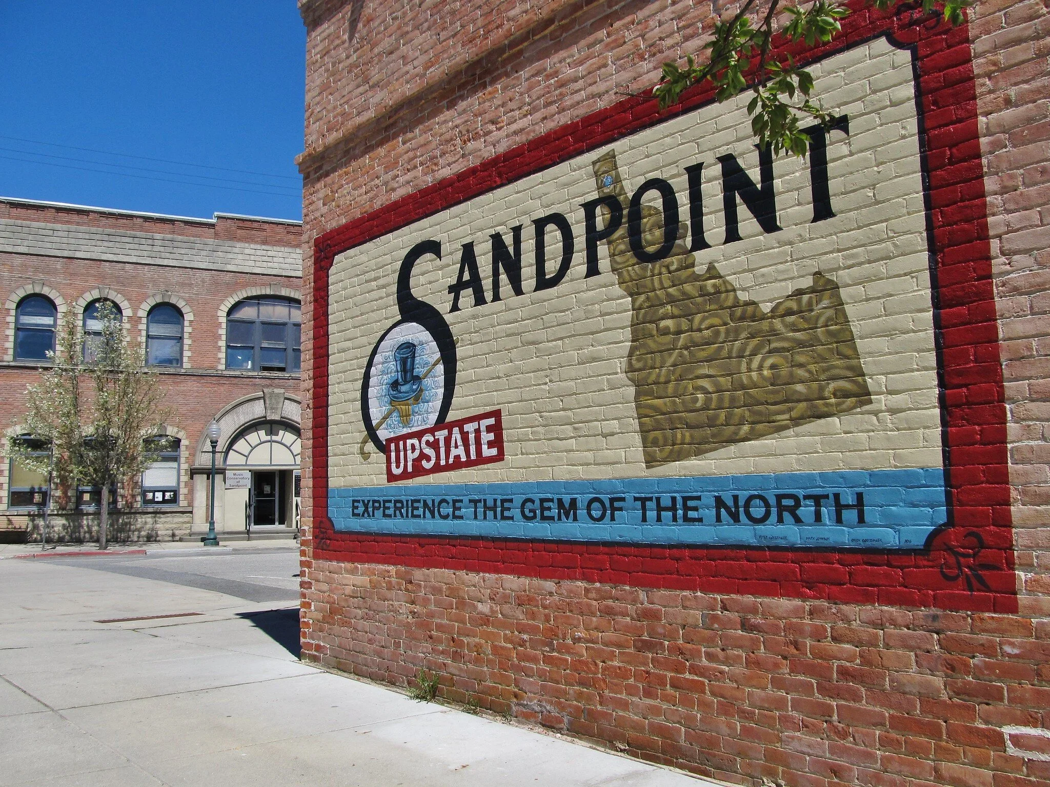 A mural on a brick wall promoting Sandpoint, Idaho, with a gold nugget, a blue and gold emblem, and the text 'Experience the gem of the North.'