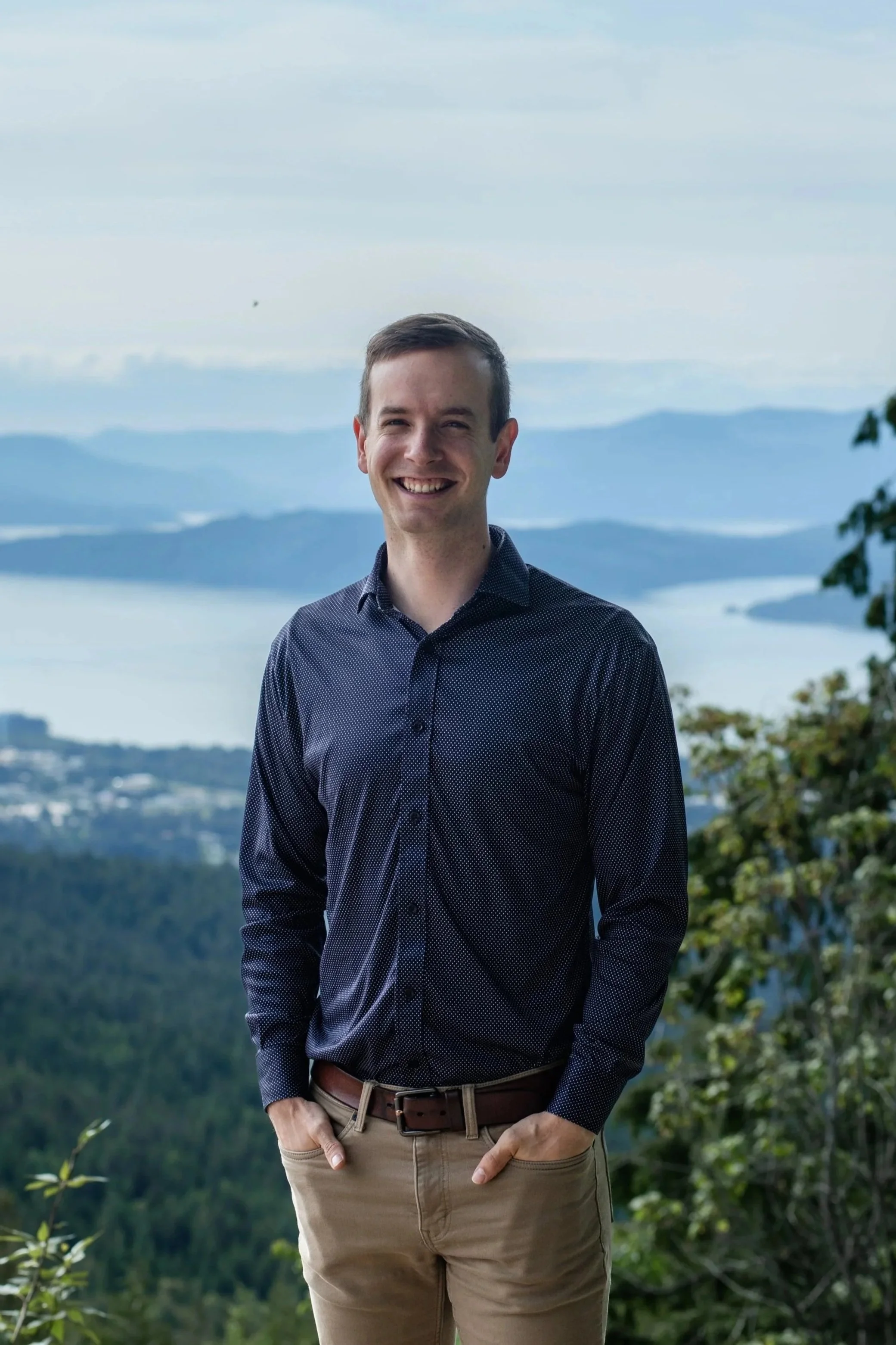 A smiling man standing outdoors with a scenic landscape of water, mountains, and greenery in the background.