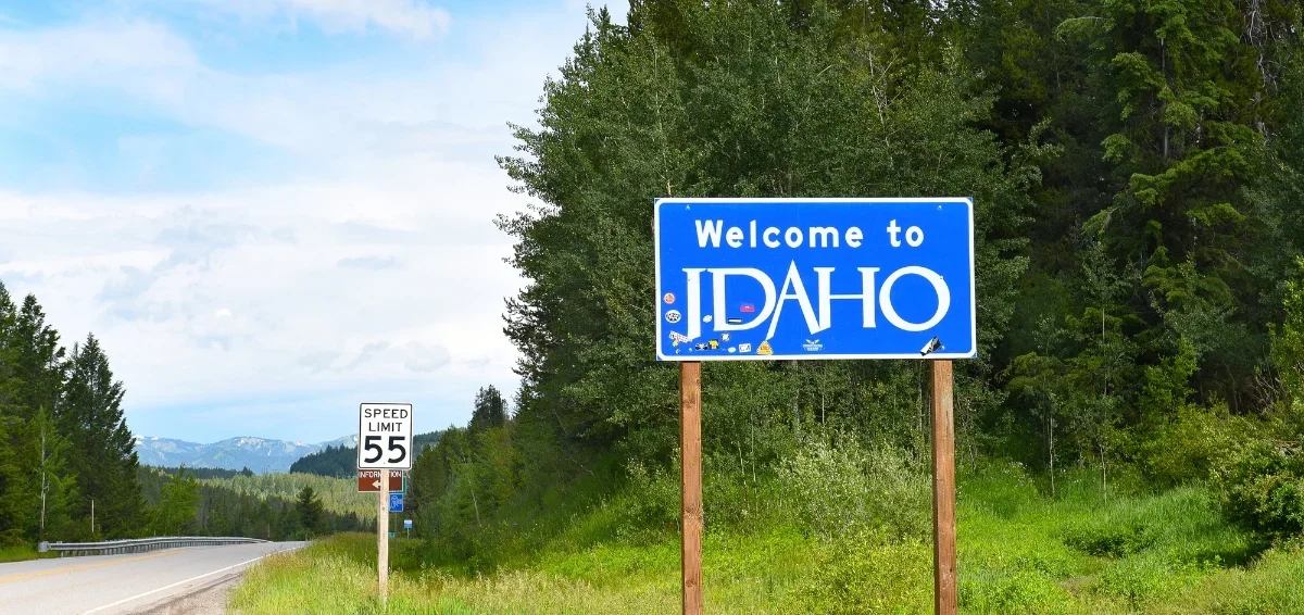 A blue sign that reads 'Welcome to Idaho' on a grassy roadside with trees and mountains in the background, and a speed limit sign showing 55 miles per hour.