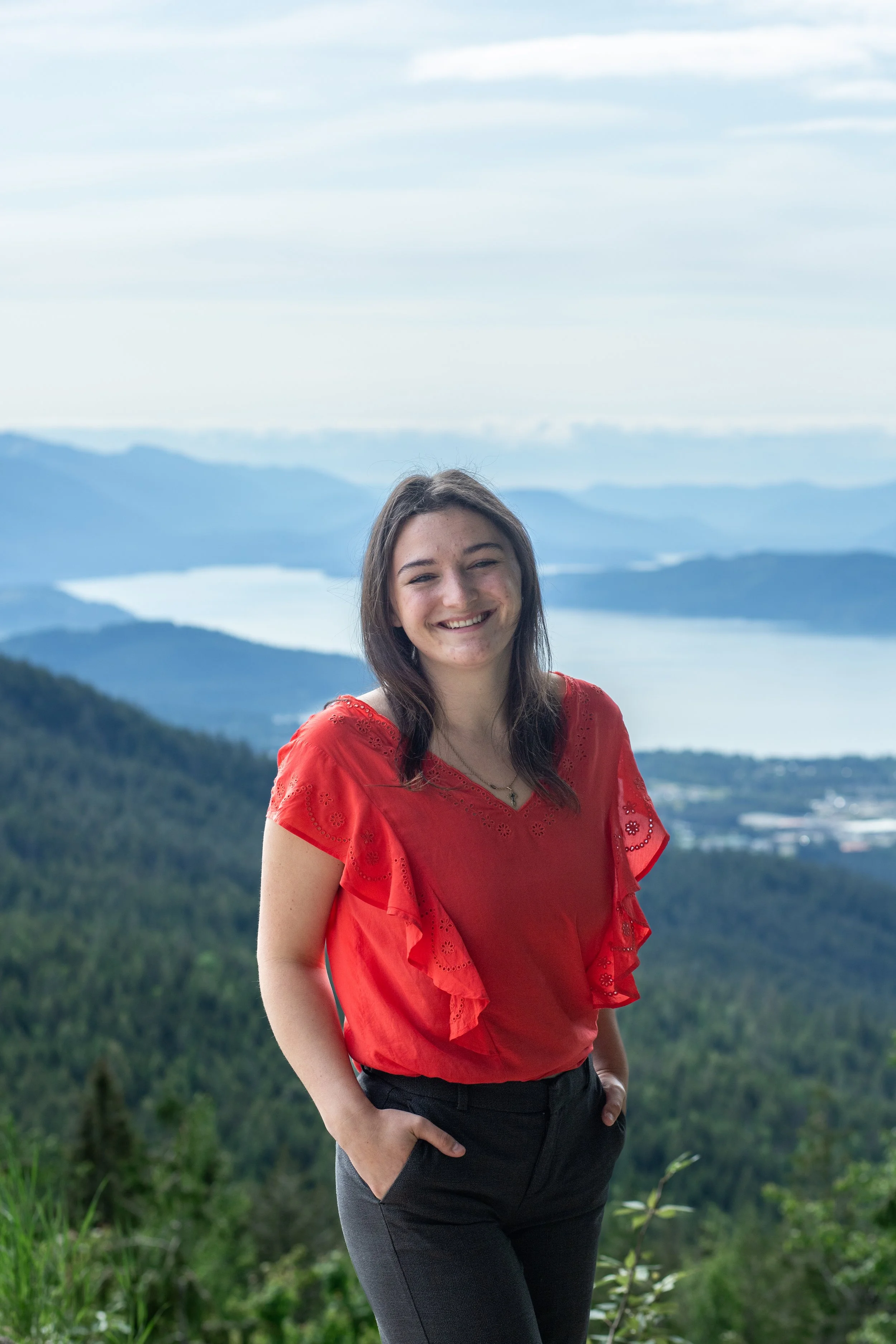 A young woman with brown hair smiling, wearing a red blouse, standing outdoors on a mountain overlooking a lake and multiple mountain ranges in the distance.