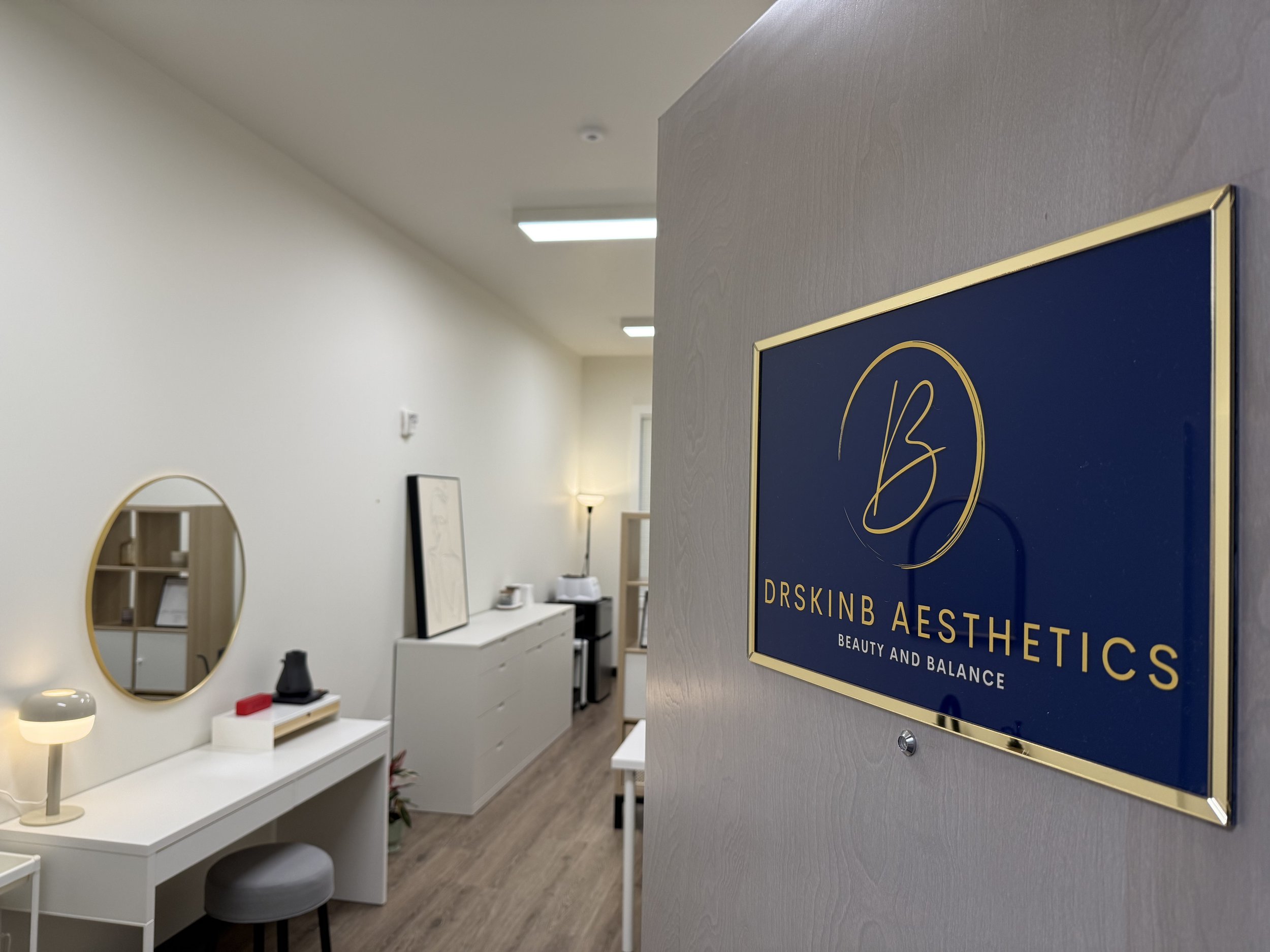 Interior of a wellness or aesthetics clinic with a sign reading 'DrSkinB Aesthetics' and the tagline 'Beauty and Balance'; minimalist decor with a mirror, a lamp, and a desk in a bright, clean space.