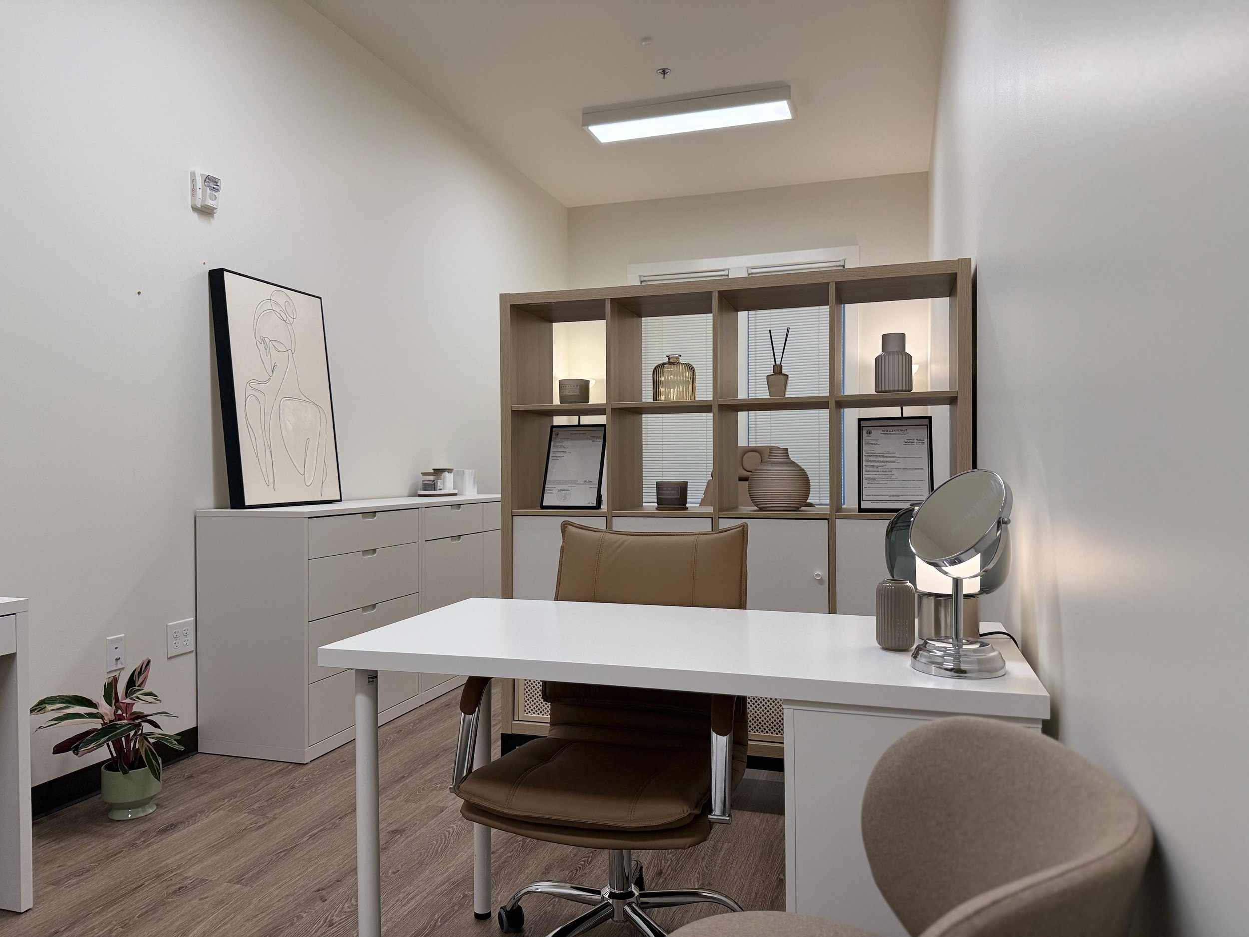 Modern office with white desk, beige chair, decorative items on a wooden shelf, and minimalist artwork on the wall.