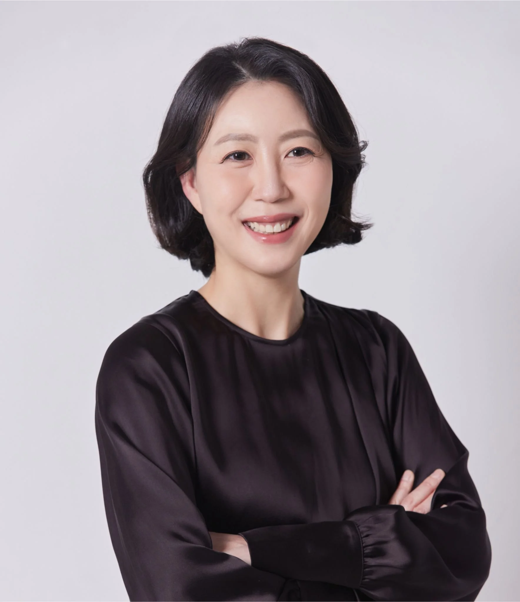 A woman with short black hair smiling, wearing a black satin blouse, standing against a plain light gray background.